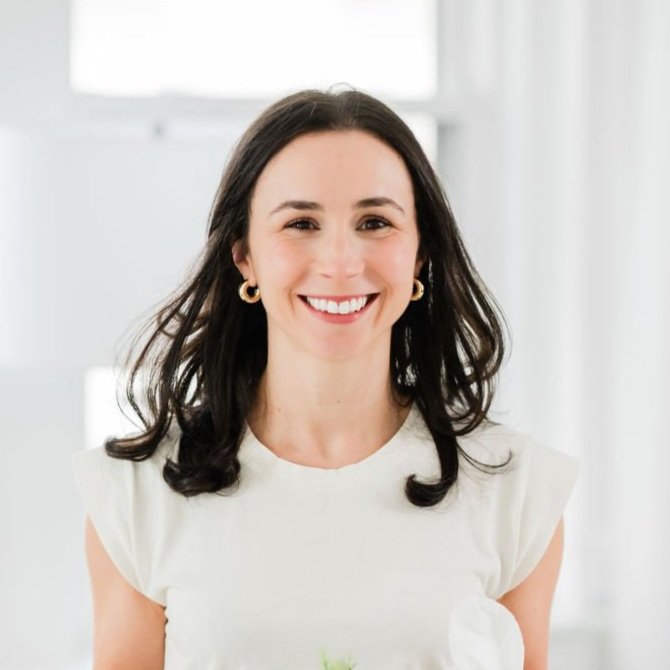 A smiling woman with shoulder-length dark hair, wearing a white shirt and gold hoop earrings, standing indoors with a bright background.
