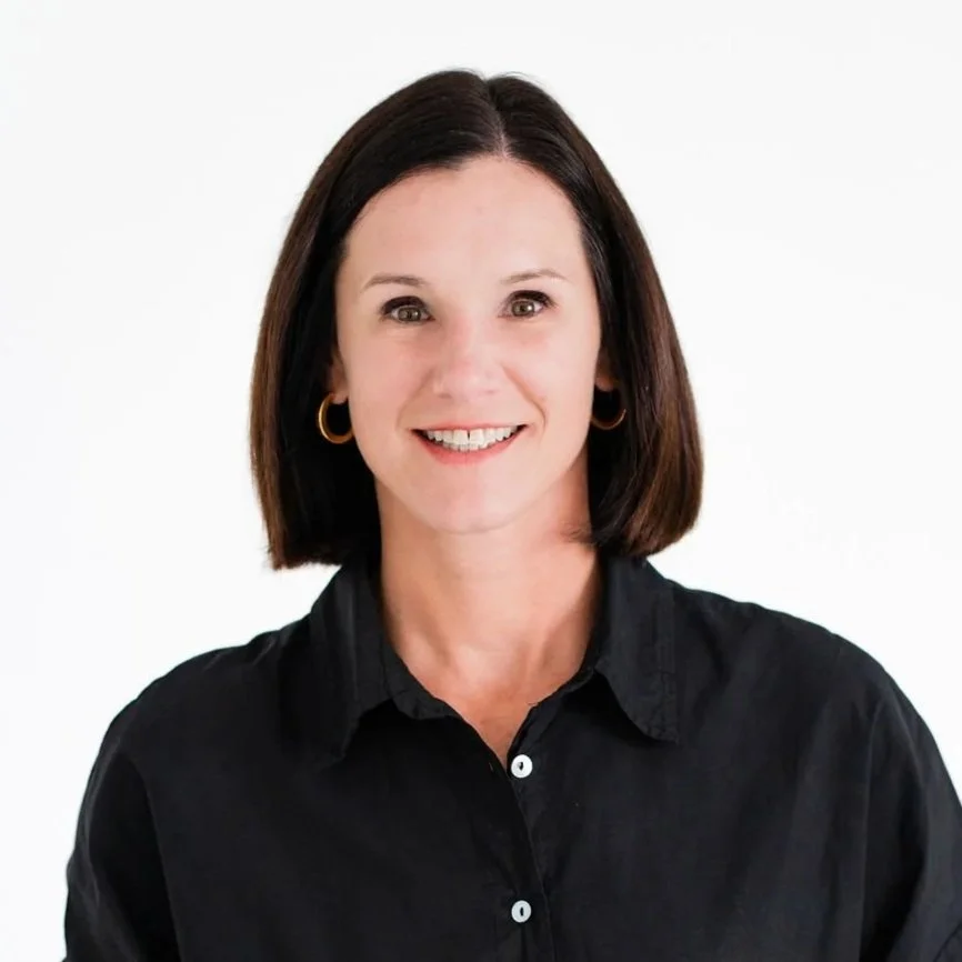 Portrait of a woman with shoulder-length dark brown hair, smiling, wearing a black button-up shirt and gold hoop earrings, against a plain white background.