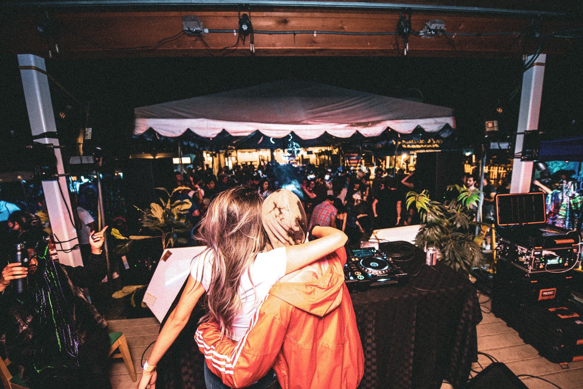 An image of DJ "episcool" and her friend hugging on stage, looking over a large crowd in the beer garden at night.