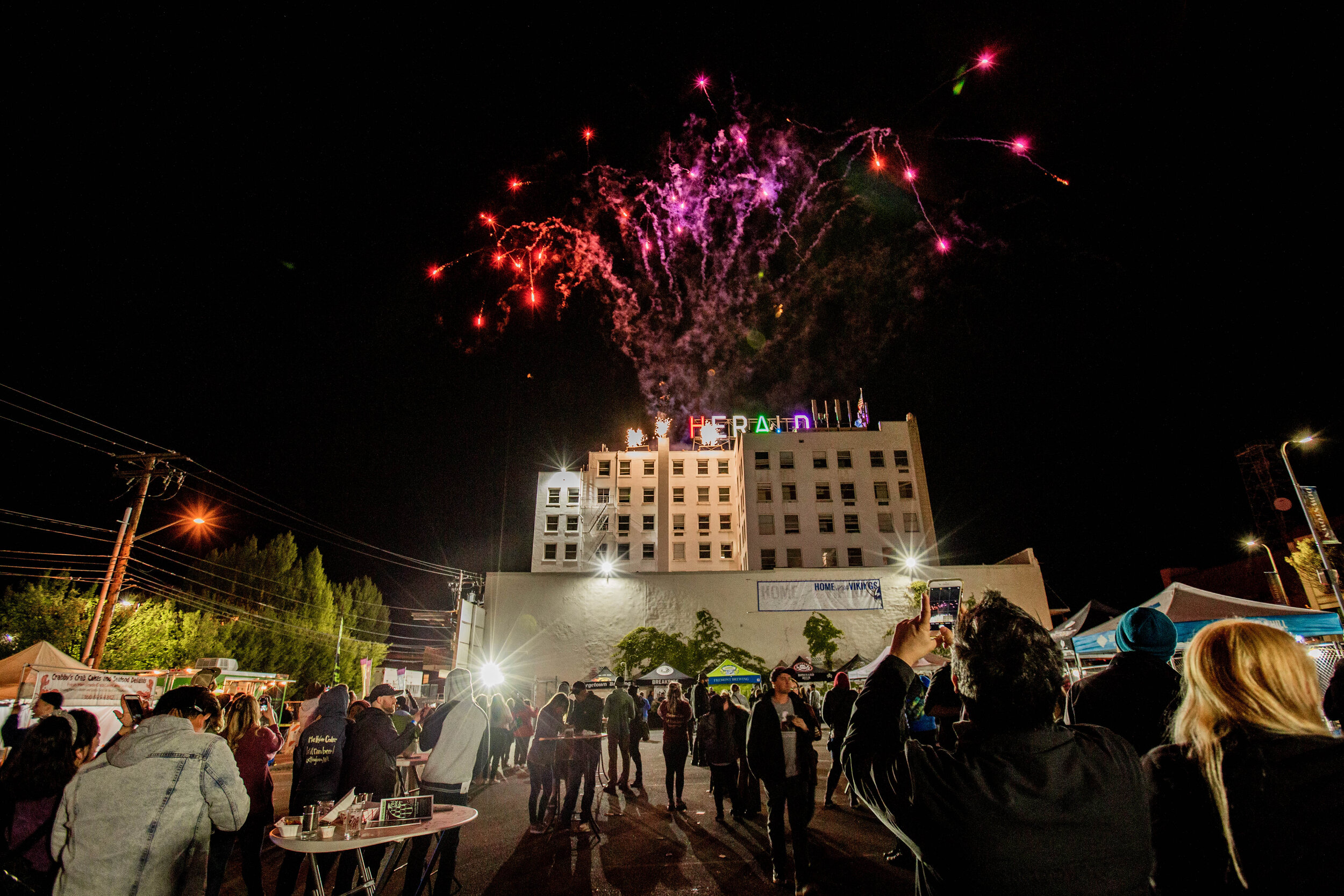 An image of a crowd gathered in Bellingham's downtown market square, watching fireworks explode over the iconic Herald sign.