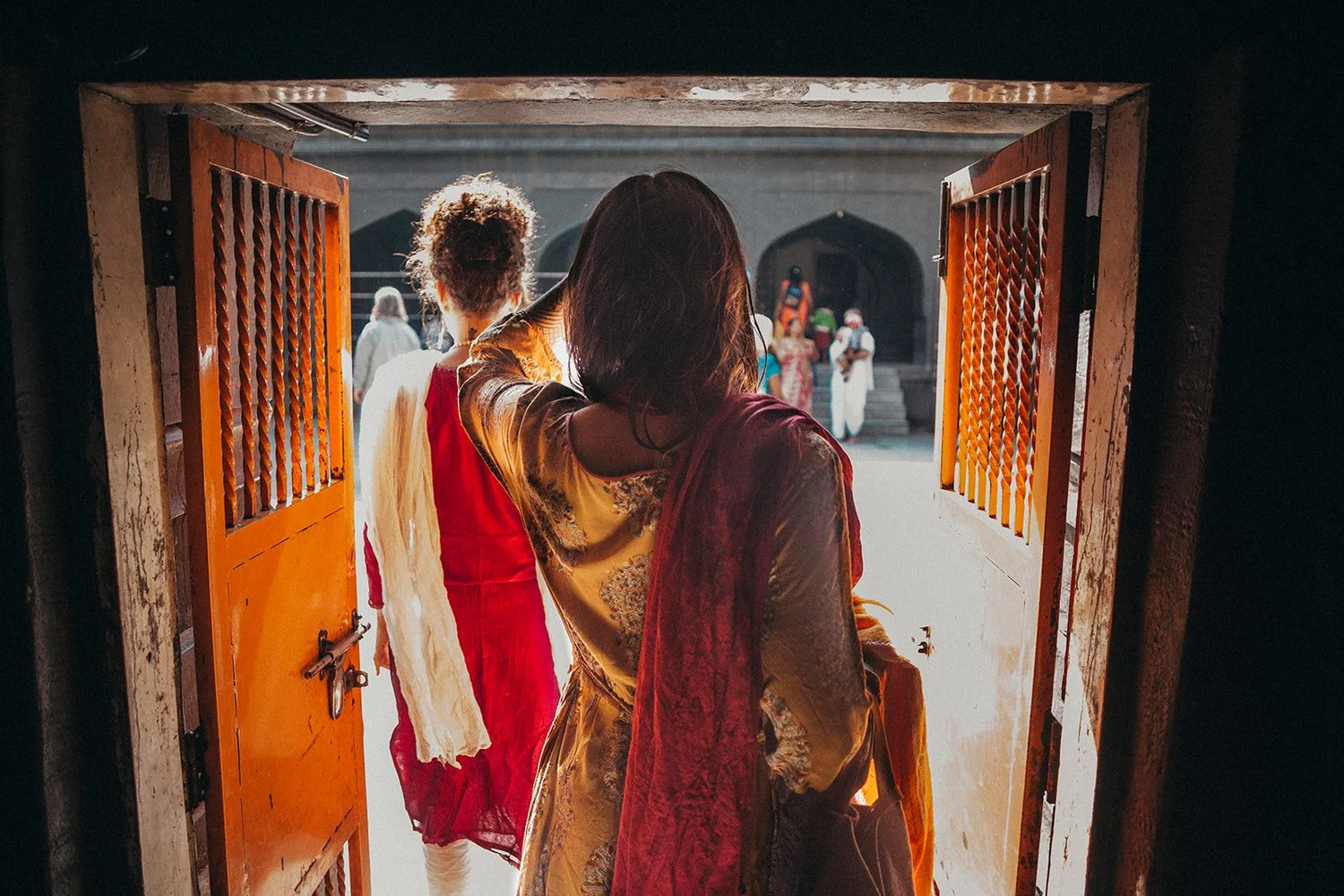 Pilgrims in red and saffron stepping through an orange temple doorway into a sunlit courtyard