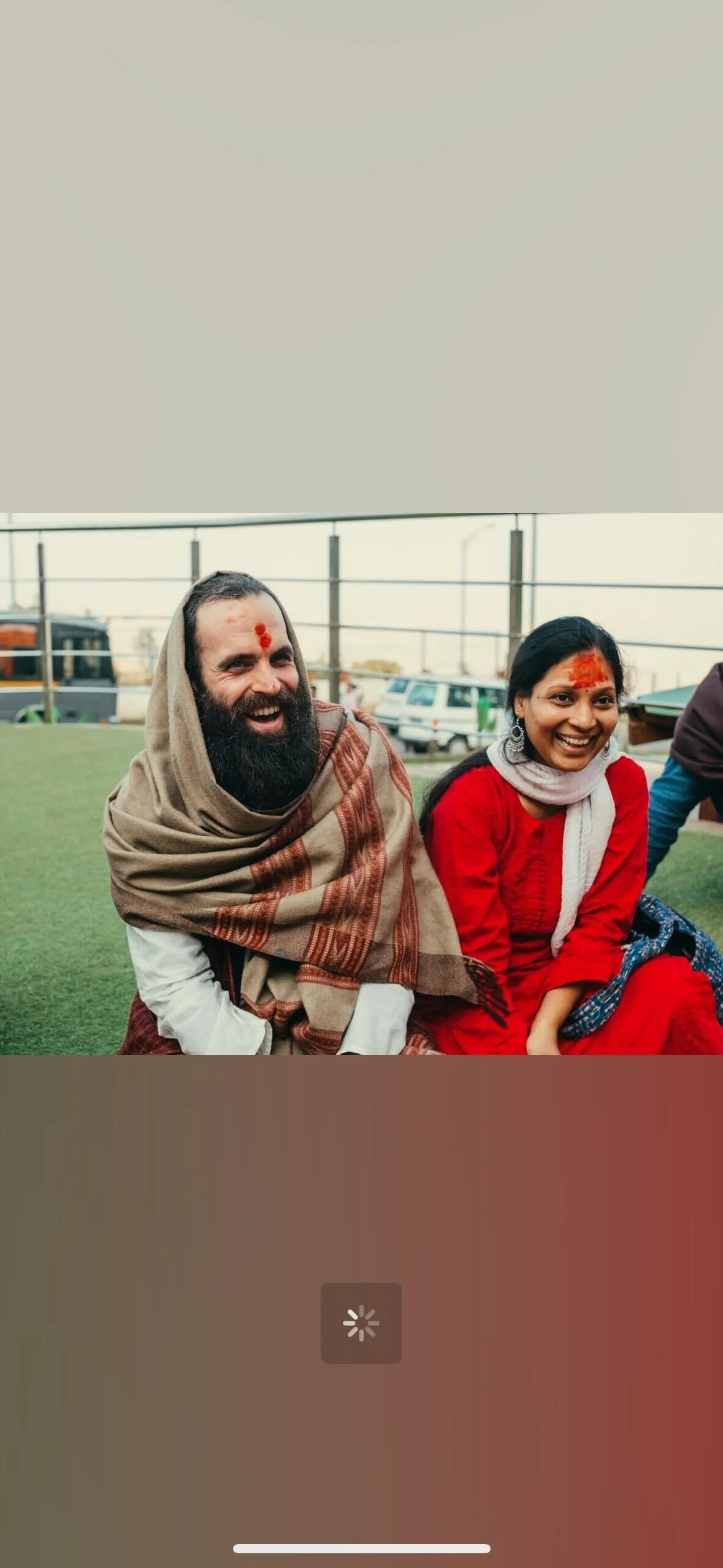Smiling man and woman sitting outdoors on green turf, with vehicles and a fence in the background, both wearing traditional South Asian attire and red forehead markings.