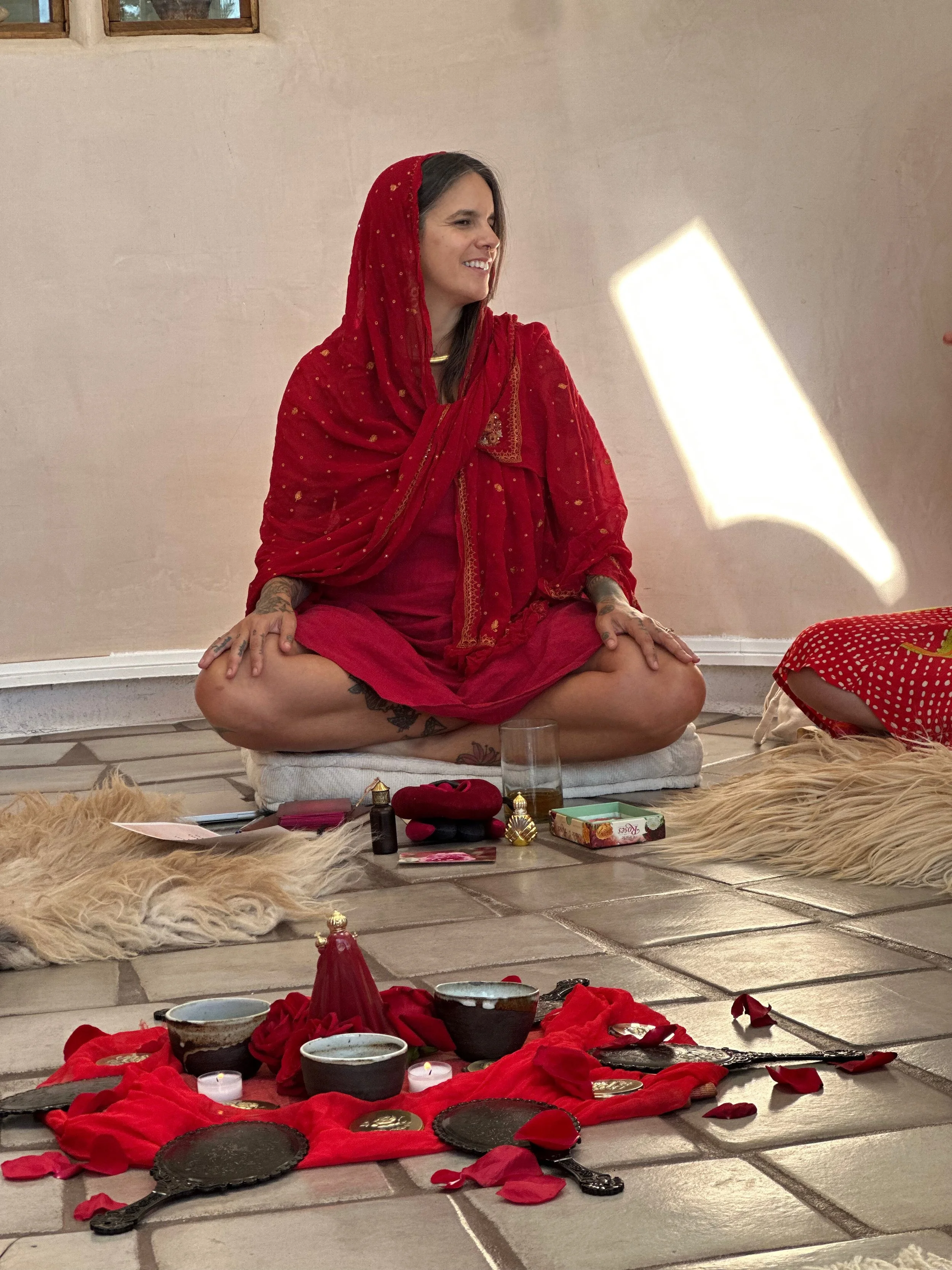 A woman dressed in a red sari sits cross-legged on a cushion on the floor, smiling. In front of her are ceremonial items including bowls, candles, and rose petals arranged on a red cloth.
