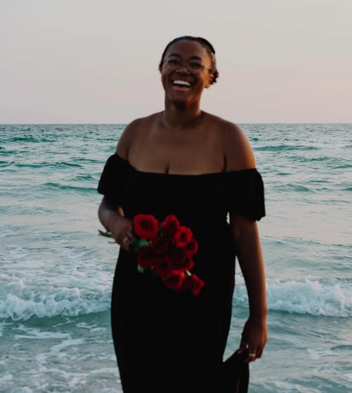 Woman smiling on the beach holding a bouquet of red flowers, wearing a black off-shoulder dress with the ocean in the background at sunset.