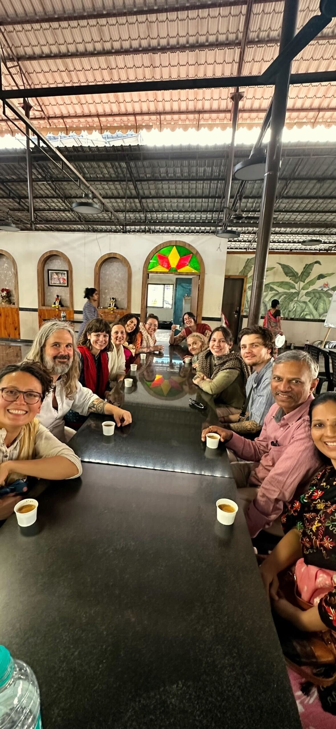A group of diverse people sitting around a long black table in a restaurant, enjoying drinks and smiling for the photo.