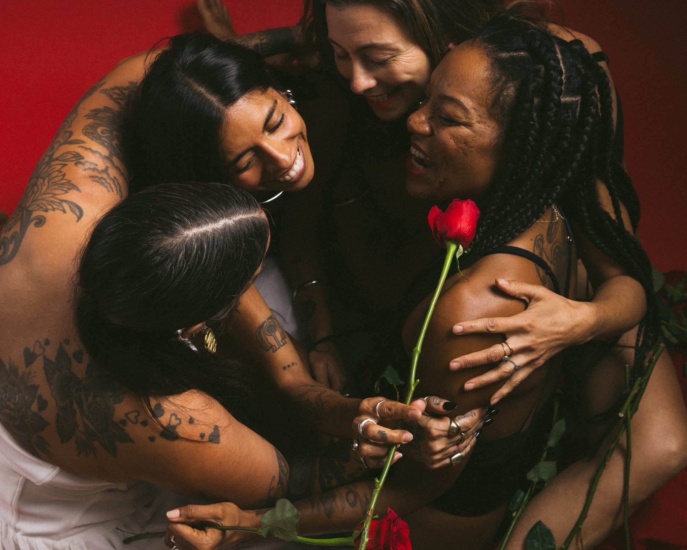 Group of five diverse women embracing and smiling, one holding a red rose, in an intimate and joyful moment against a red background.