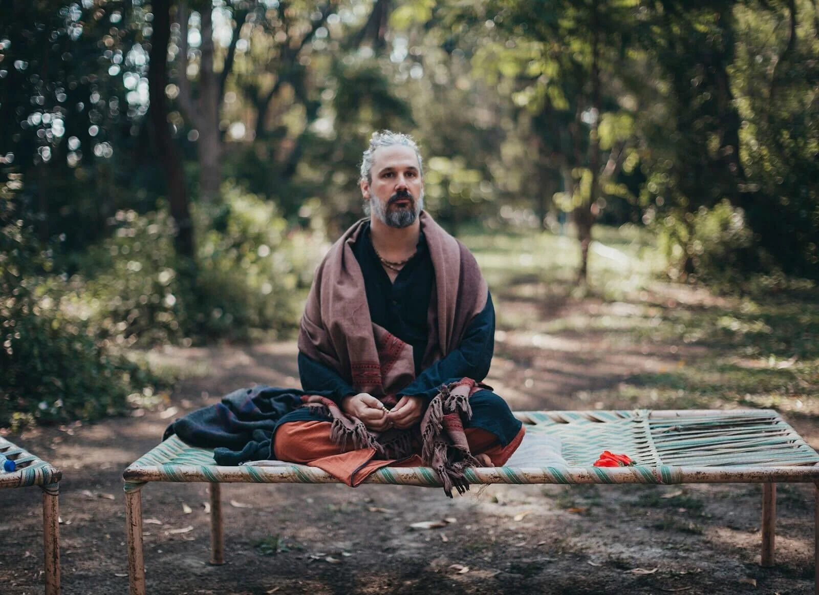 A man with gray hair and beard sitting cross-legged on a woven outdoor bed in a forest, wearing a dark shirt, orange pants, and a brown shawl, with trees and greenery in the background.