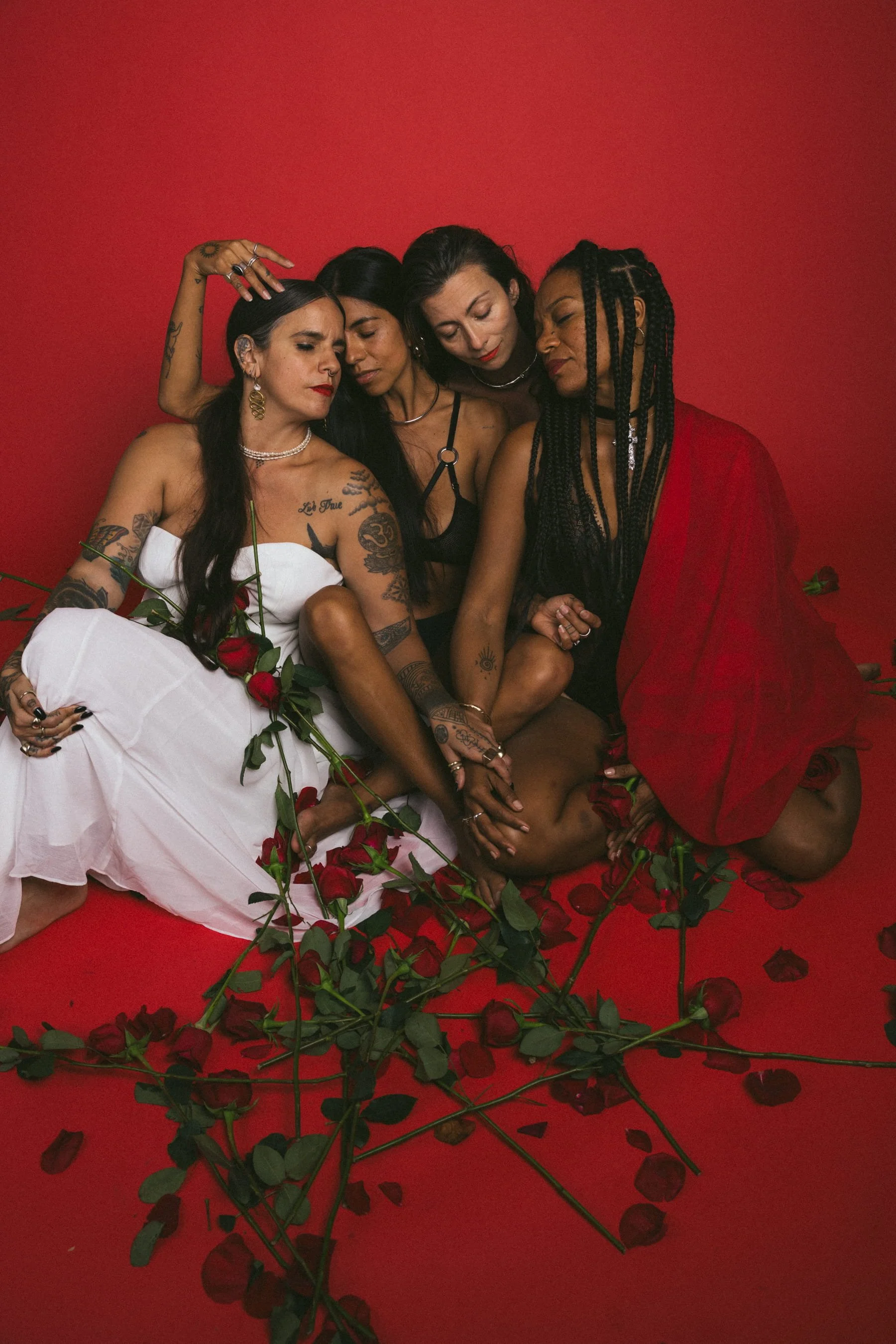 Four women sitting on a red floor with scattered rose petals and roses, dressed in black, white, and red outfits, with a red background, showing a sense of intimacy and connection.