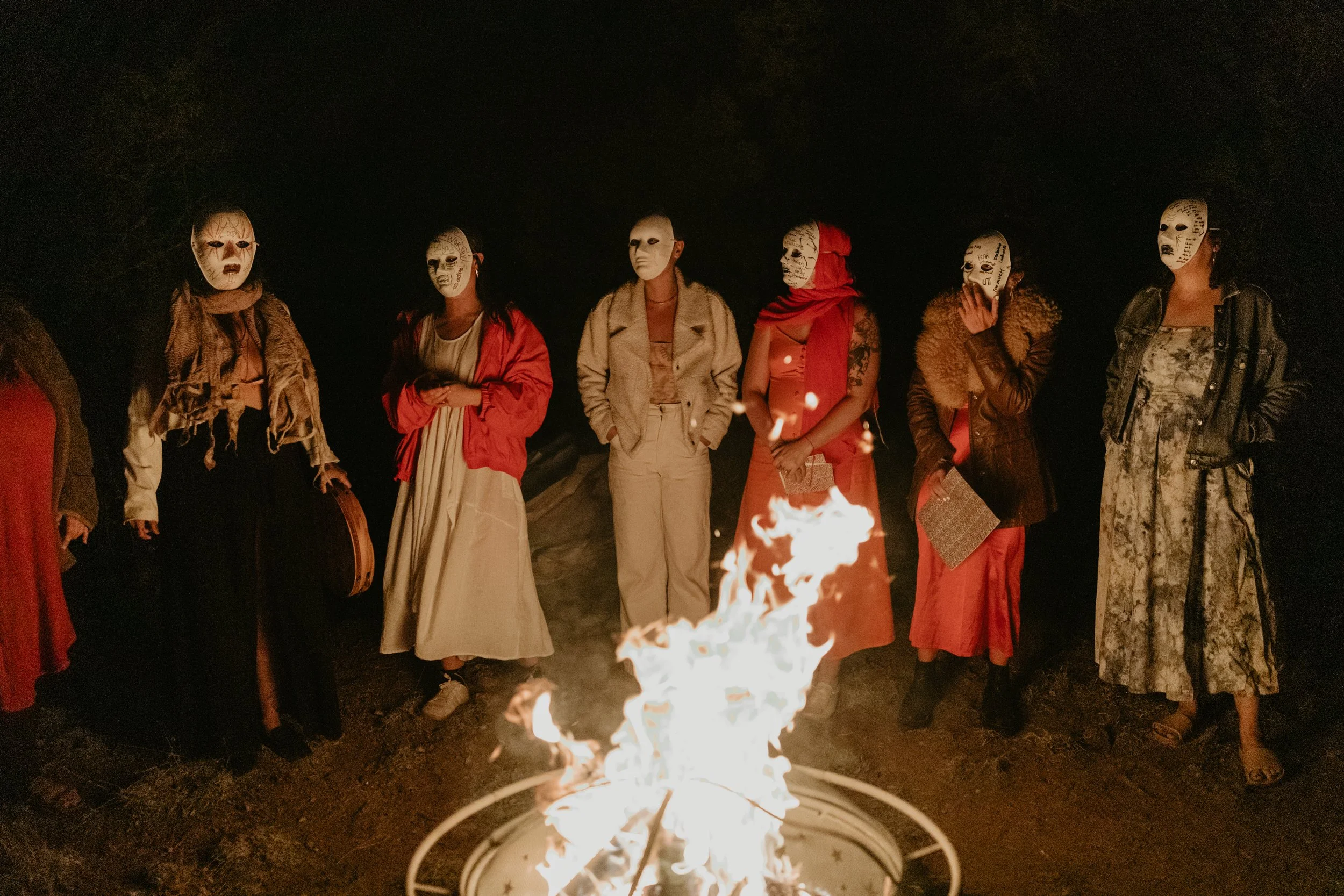 Group of people standing around a campfire at night, all wearing white masks with black markings, dressed in casual and stylish clothing.