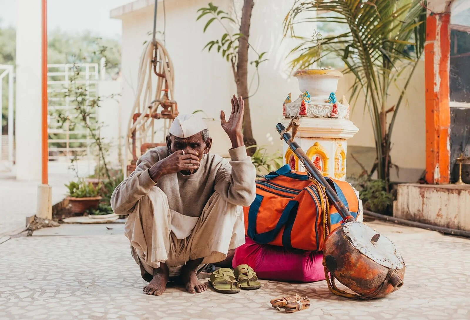An elderly man squats on a patterned tiled floor, wearing beige clothes and a white cap, holding a cup to his mouth with one hand and raising the other hand. Next to him are a pair of sandals, an orange and blue bag, a pink cushion, a drum, and a wal