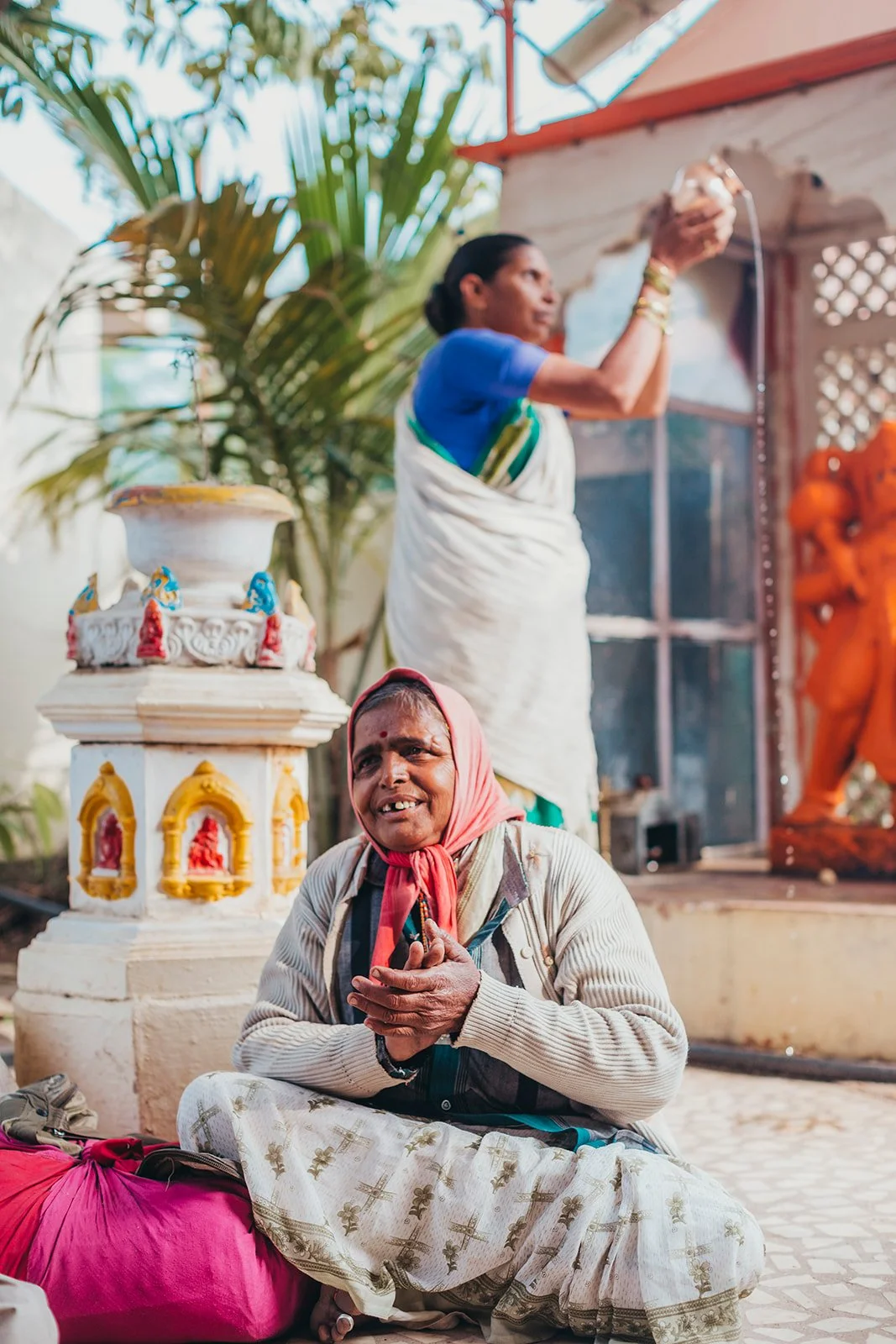 An elderly woman seated beside a small shrine, hands pressed together in prayer