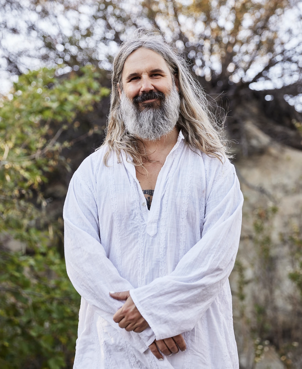 A man with long gray hair and a beard stands outdoors in front of trees and bushes, wearing a loose white shirt with sleeves rolled up, smiling at the camera.