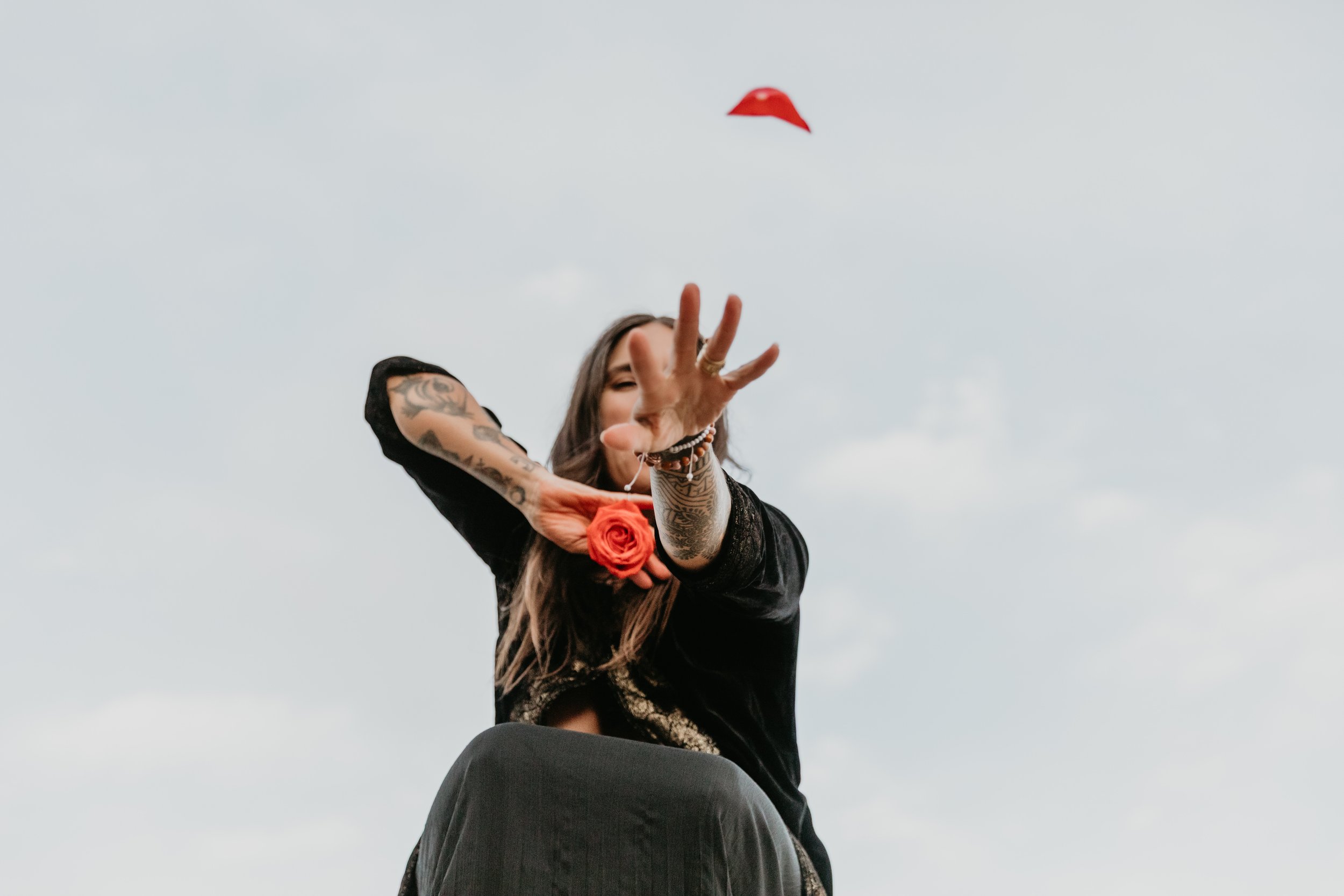 A woman with tattoos and long hair is throwing a red rose and a red paper hat into the air during daytime