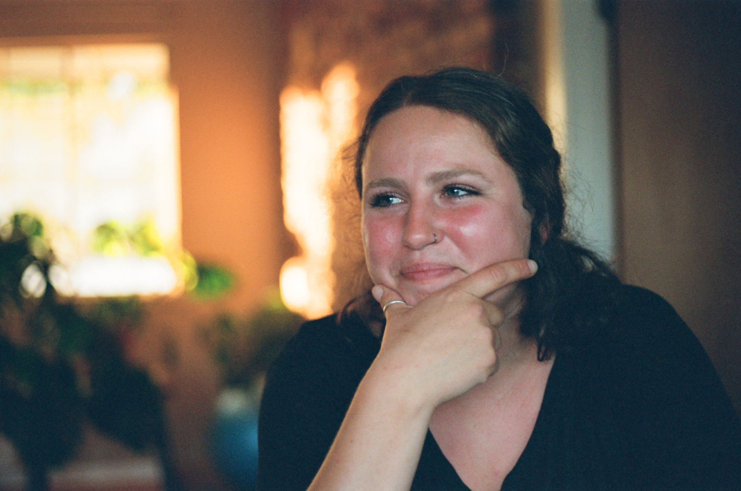 Young woman with dark hair, light skin, and a nose ring sitting in a cozy indoor setting, resting her chin on her hand, with a thoughtful or whimsical expression.