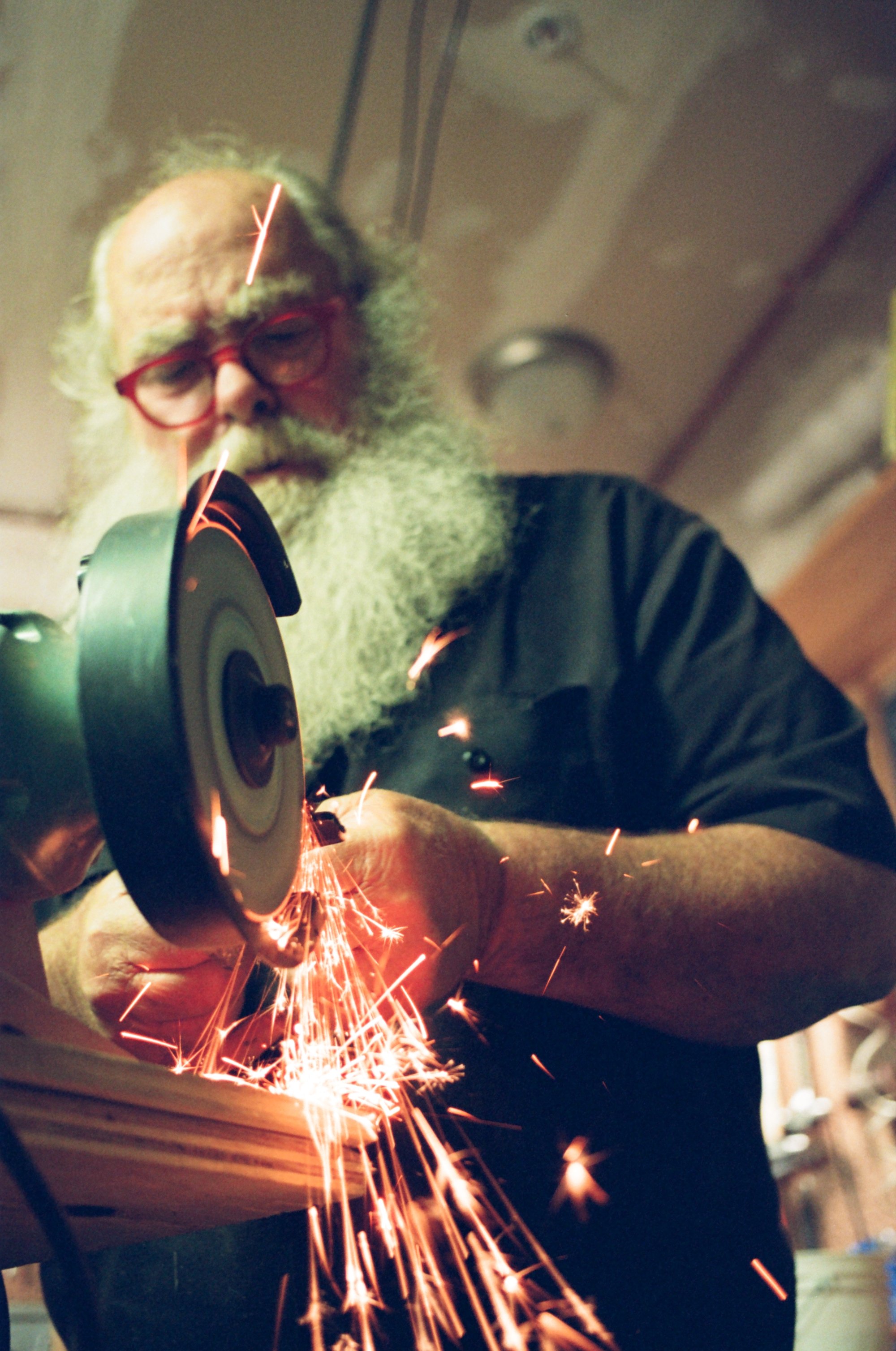 An elderly man with a white beard and glasses uses a power grinder, creating sparks, in a workshop setting.