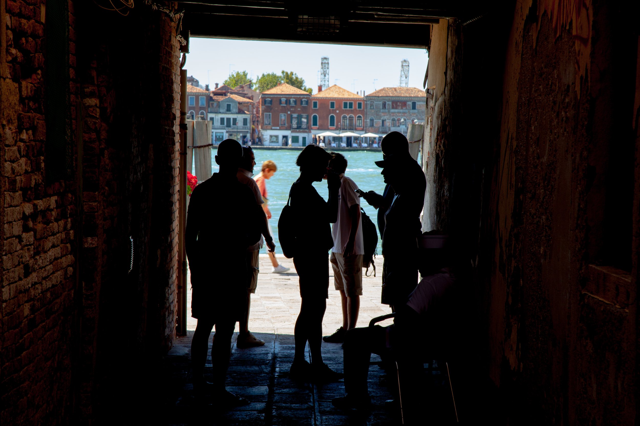Silhouetted group of people standing in a passageway opening to a waterfront scene with colorful buildings across the water in Venice, Italy.