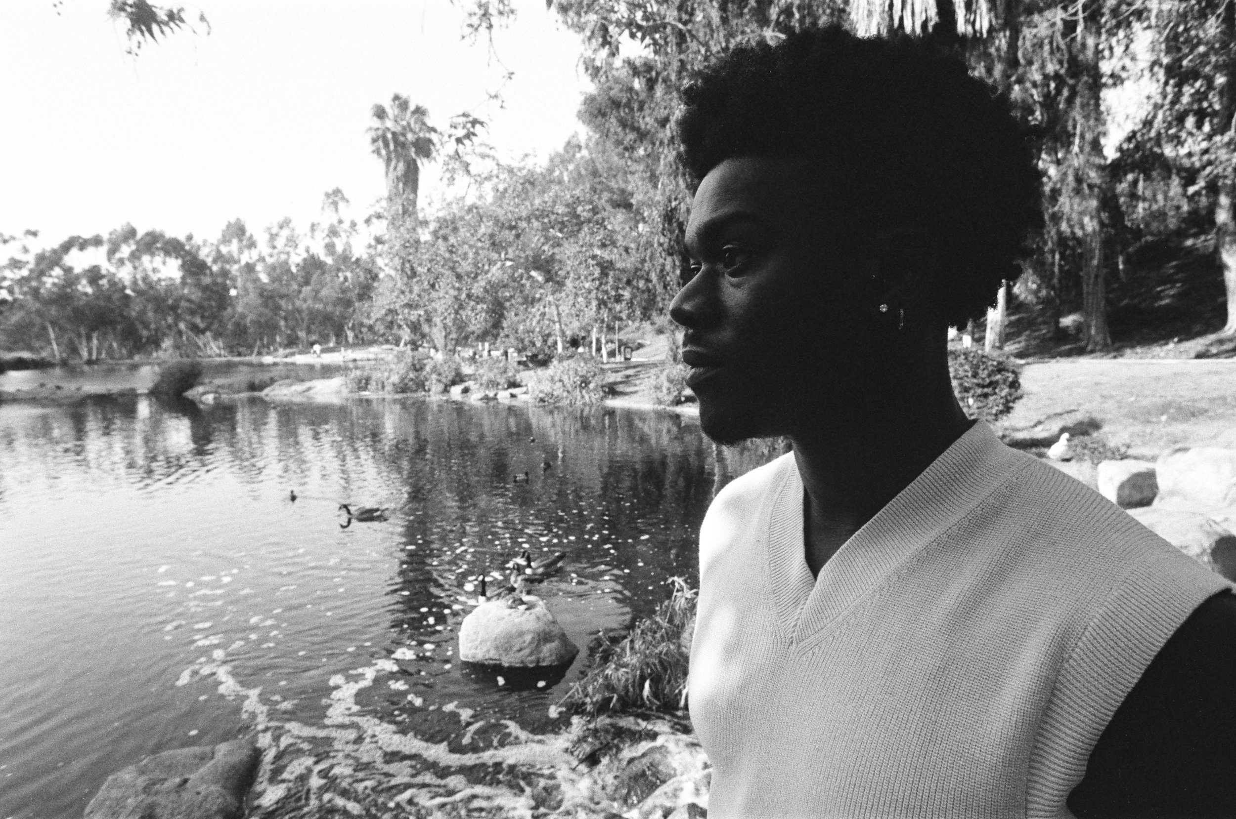 A black-and-white photo of a young man with short, natural hair standing near a body of water in a park, with trees and ducks in the background.