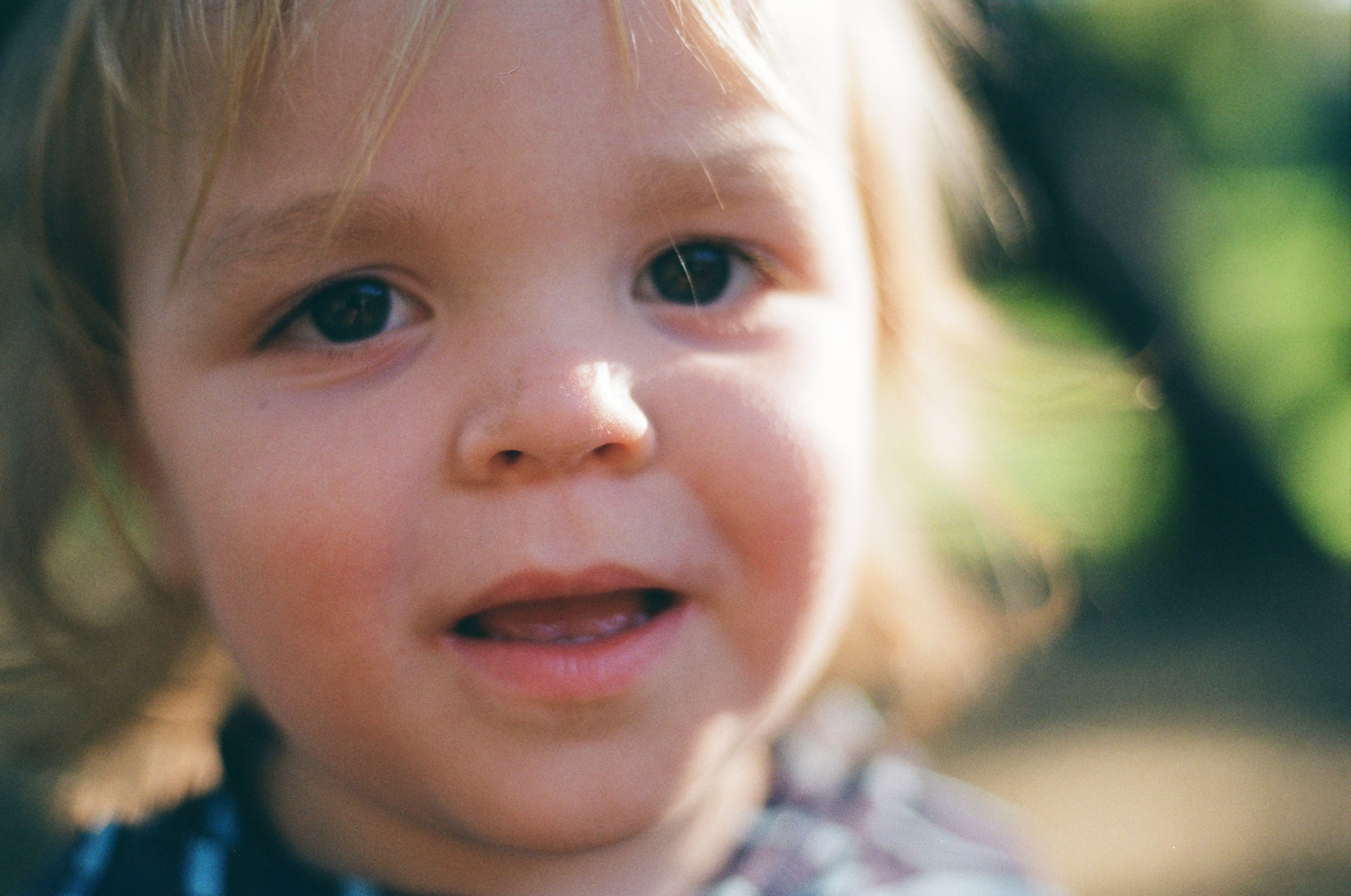 Close-up of a young child with light brown hair and blue eyes, outdoors in natural light, with a blurred green background.