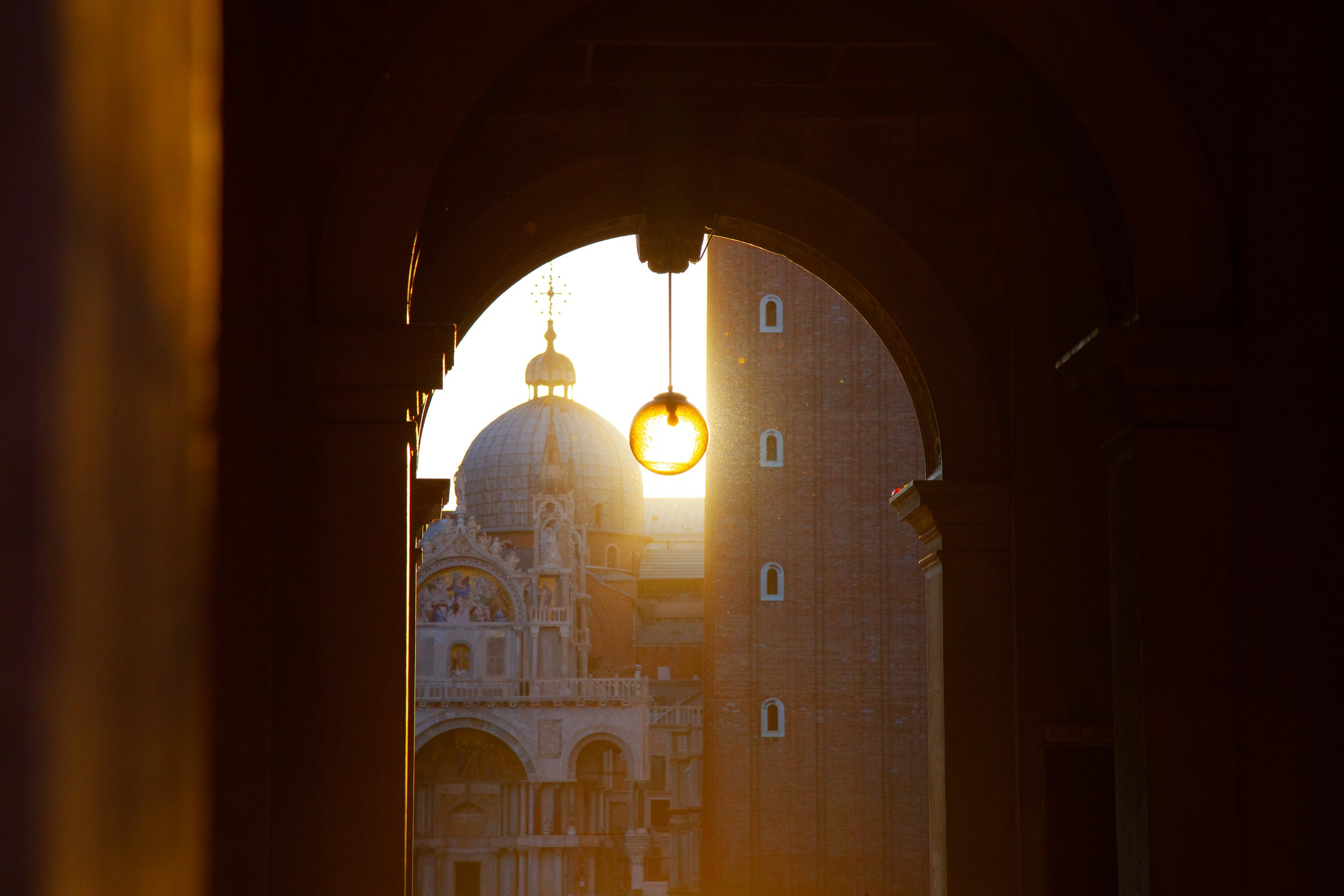 View of a domed church through an arched window, with a decorative chain overhead and a brick tower on the right, during sunset.