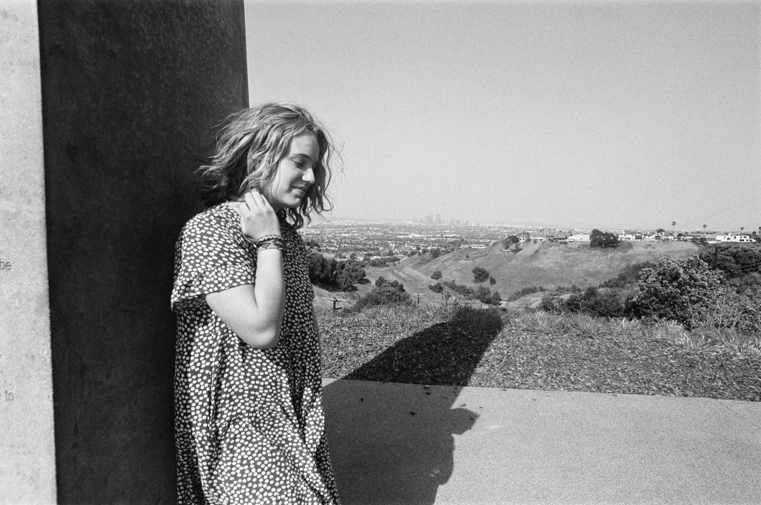 A young woman with curly hair wearing a polka dot dress is leaning against a wall outdoors, with a scenic view of hills and city in the background on a sunny day.