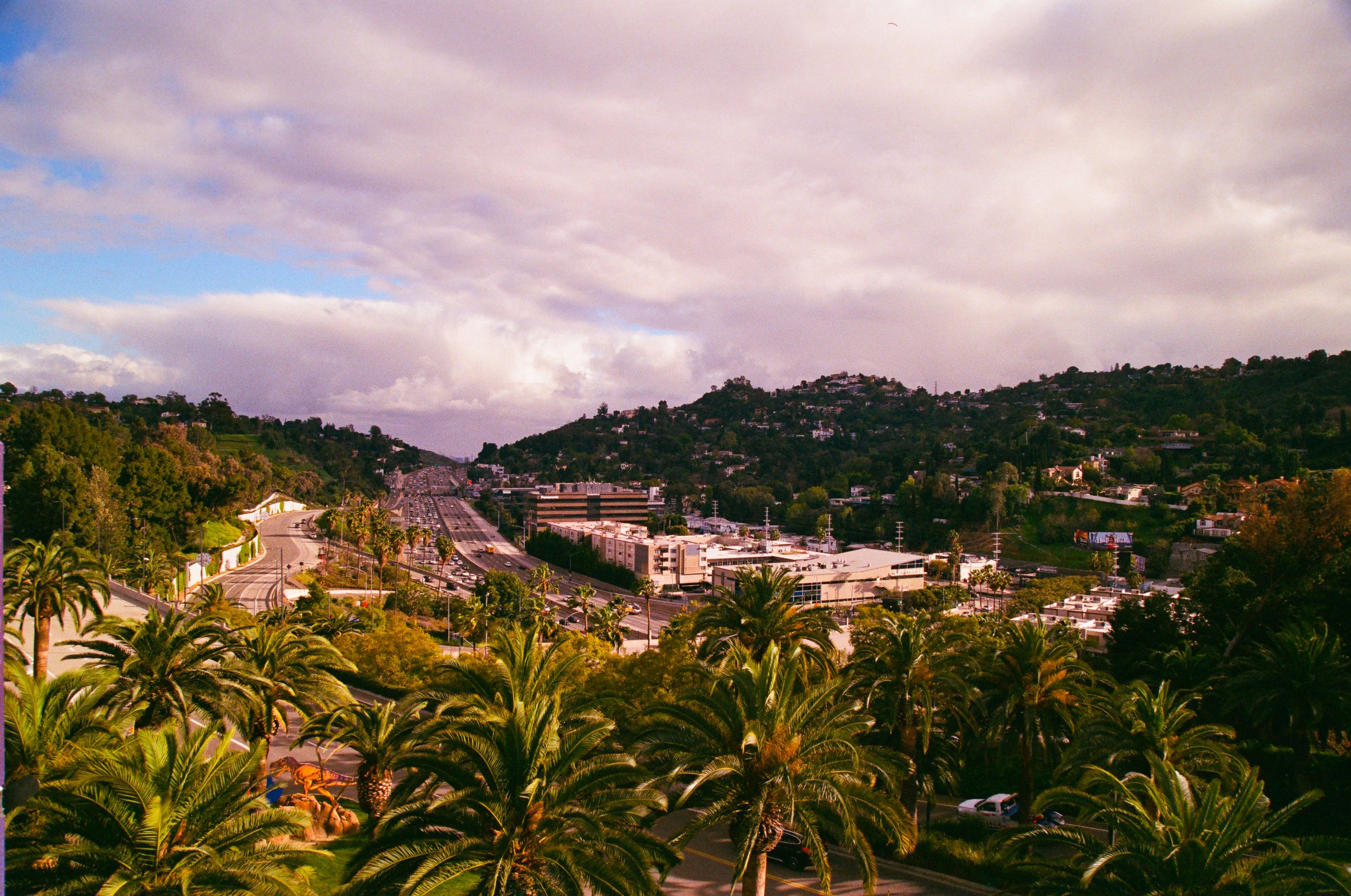 A view of a cityscape with a highway, surrounded by lush green palm trees and hills under a partly cloudy sky.
