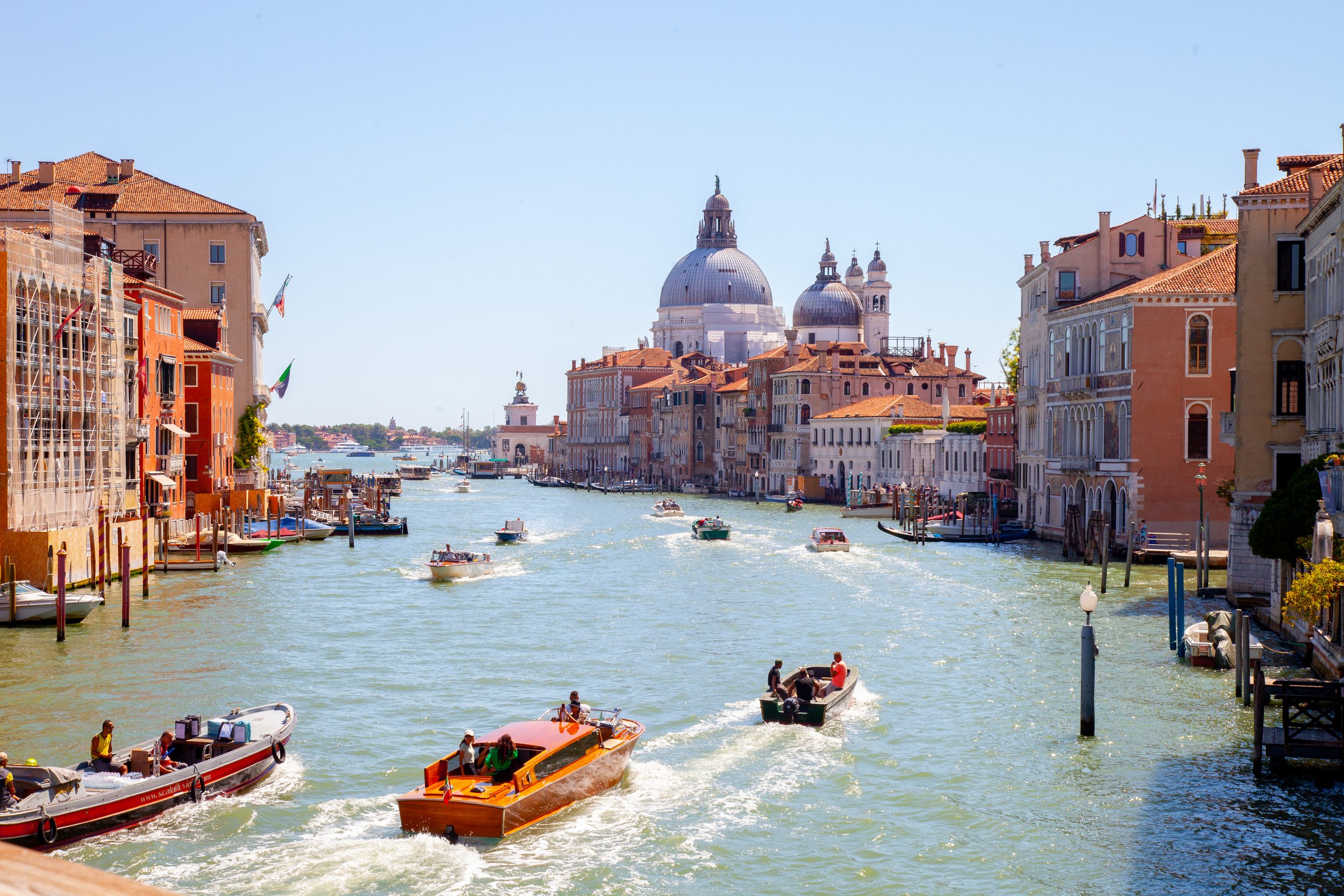 Venetian canal with boats and colorful historic buildings, with the church of Santa Maria della Salute in the background, under a clear blue sky.
