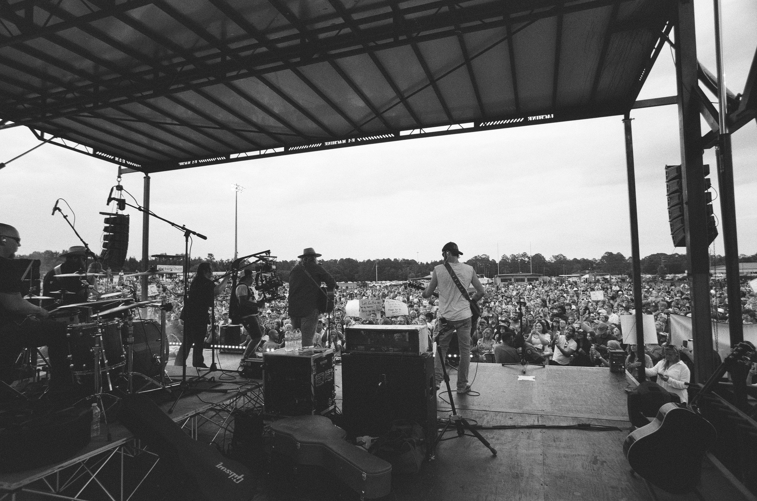 Black and white photo of a large outdoor concert stage with musicians performing, facing a big crowd of people holding signs and wearing hats, with some trees and structures in the background.