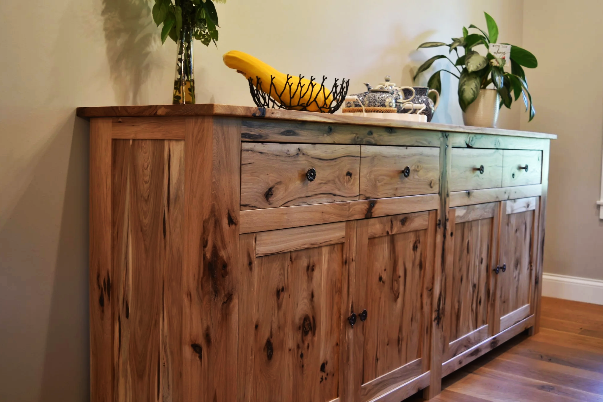 Wooden sideboard with plants, teapot, and banana on top, in a room with beige walls and hardwood floor.