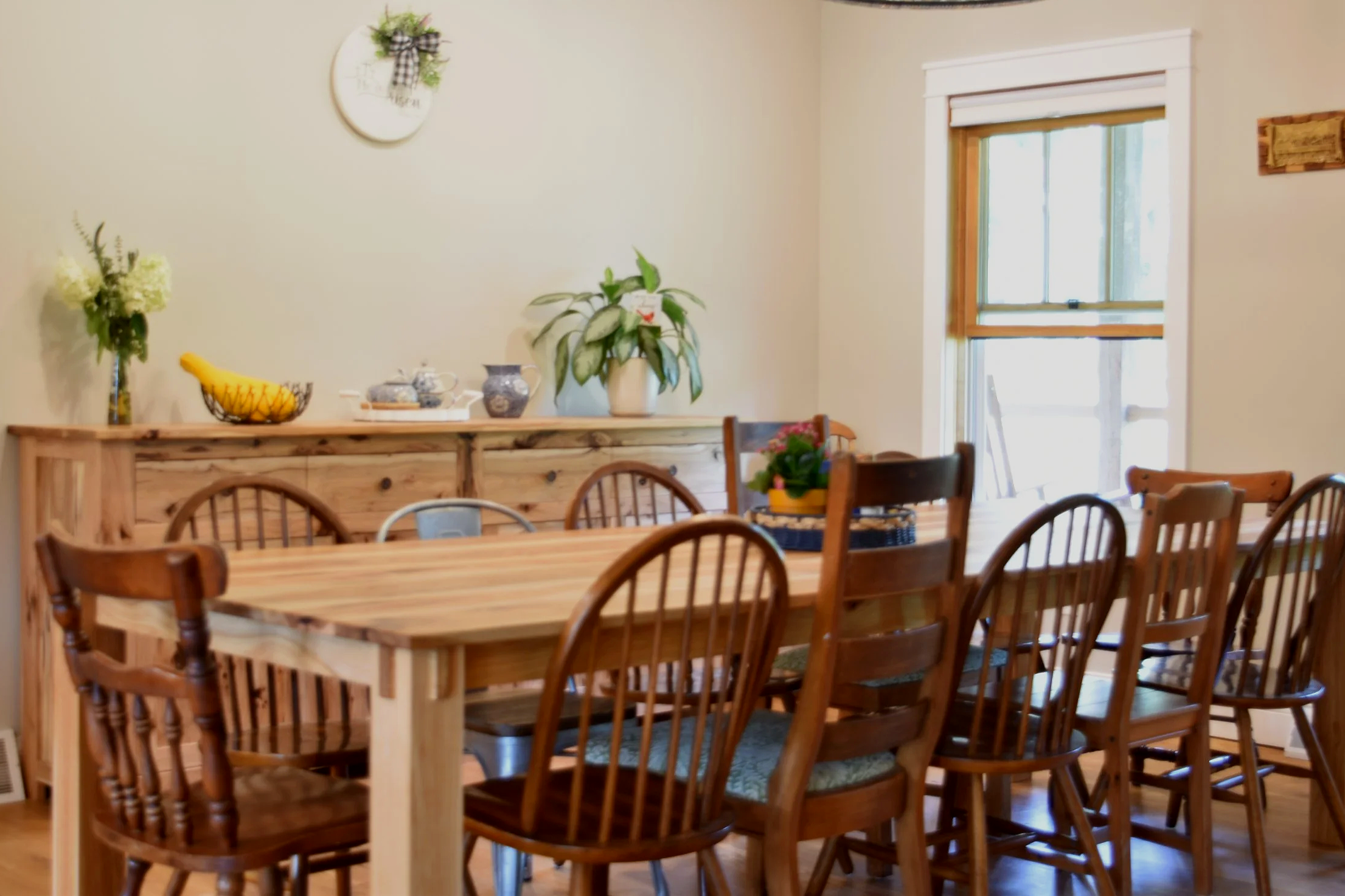 Dining room with wooden table and chairs, a sideboard with decorative items, a window with wooden trim, and various plants and decorations on the wall and sideboard.