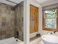 Bathroom with a shower area featuring beige tiles and a brass shower faucet, next to a window with wooden shutters and a round white sink.