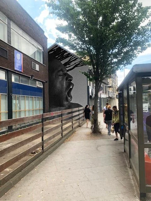 Urban street scene featuring a black and white mural of a person with an open mouth, possibly shouting or singing, painted on the side of a building. Several people are walking past on the sidewalk, and there's a bus stop on the right. Trees and mode