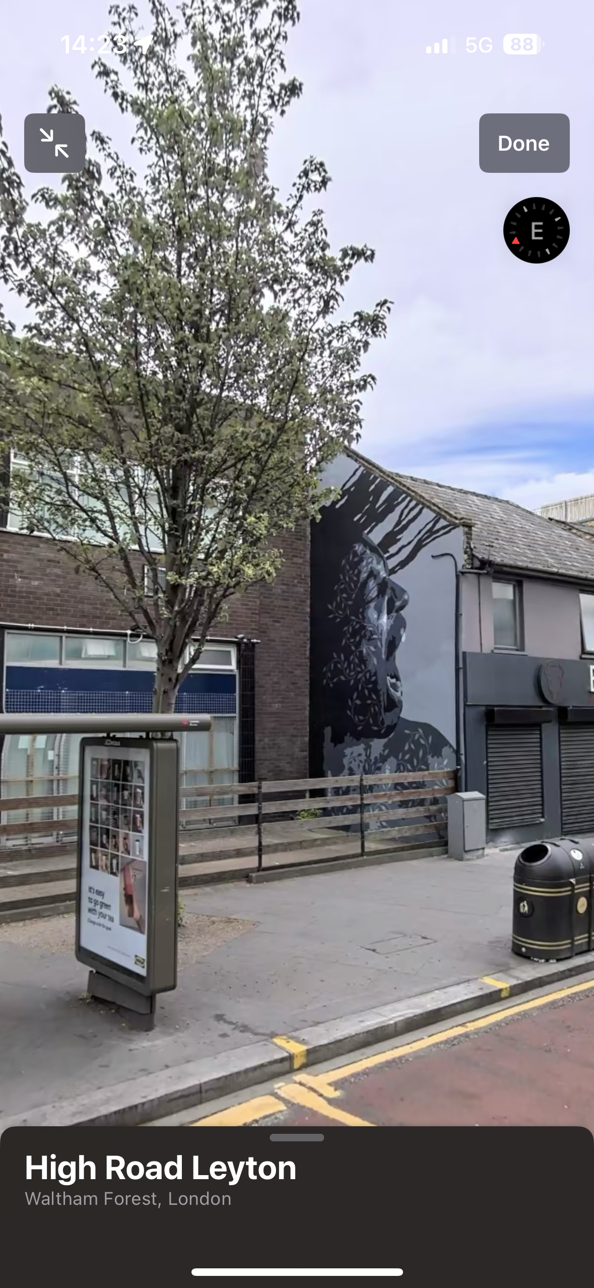Street scene in Leyton, London showing a bus stop, tree, mural on a building, and garbage bin.
