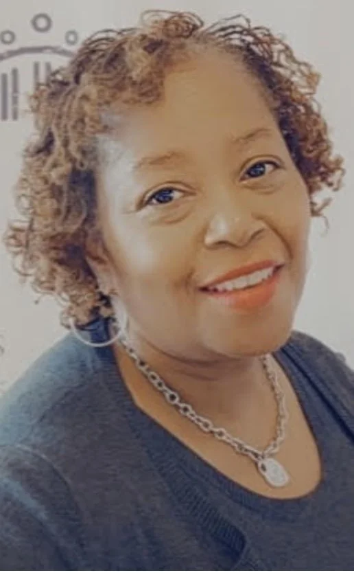 A smiling woman with curly, light brown hair, wearing a black top, a silver chain necklace, and hoop earrings. A clock is visible in the background.