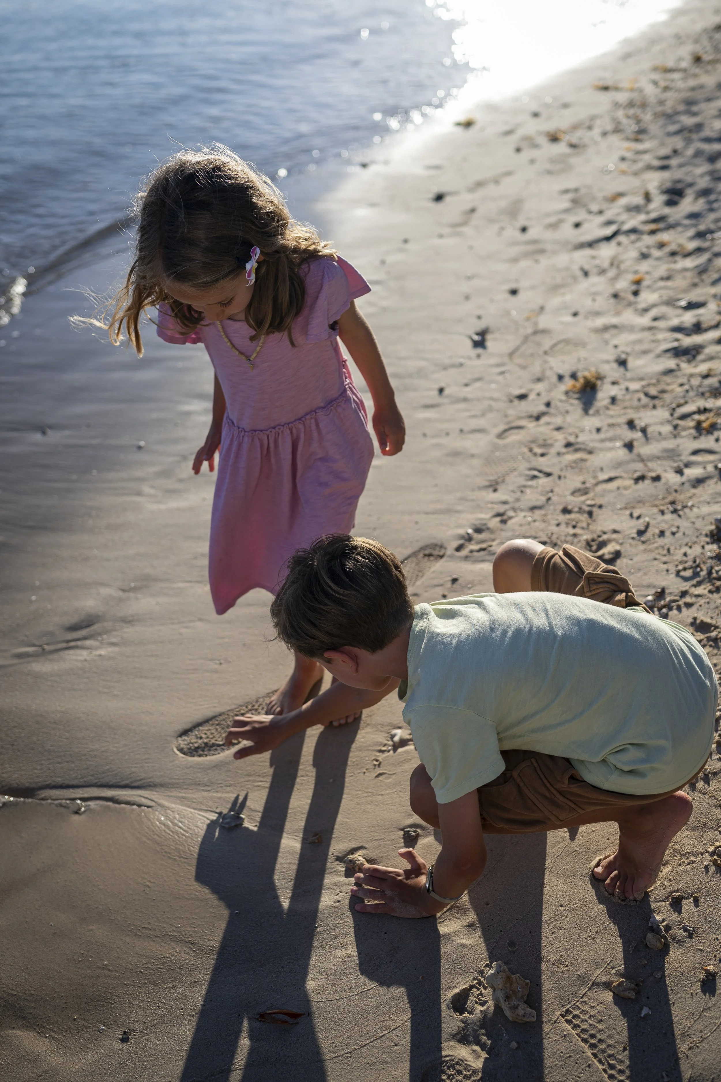 Two children, a girl and a boy, are playing on the beach near the water. The girl is standing and looking down, while the boy is crouched down, touching the sand and rocks.