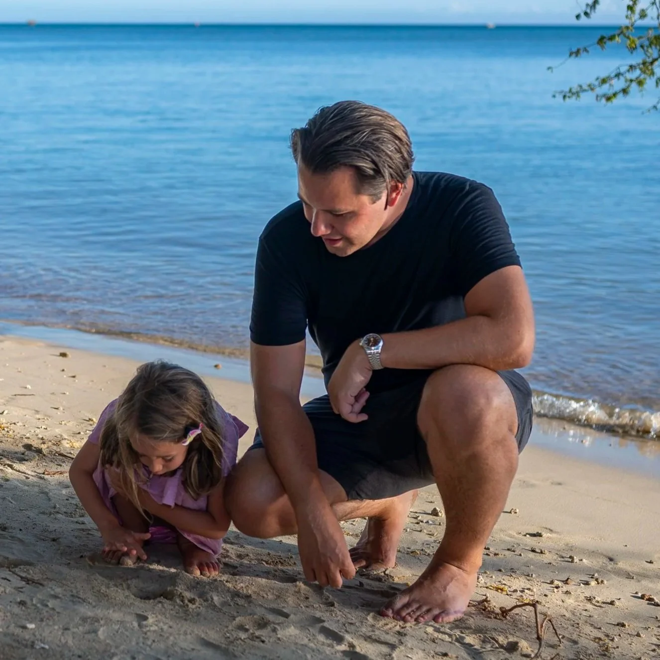 A man and a young girl are crouching on a sandy beach by the water, looking at the sand closely. The man is wearing a black t-shirt, shorts, and a watch, while the girl is wearing a purple dress. The scene takes place during the day with a calm body of water in the background.