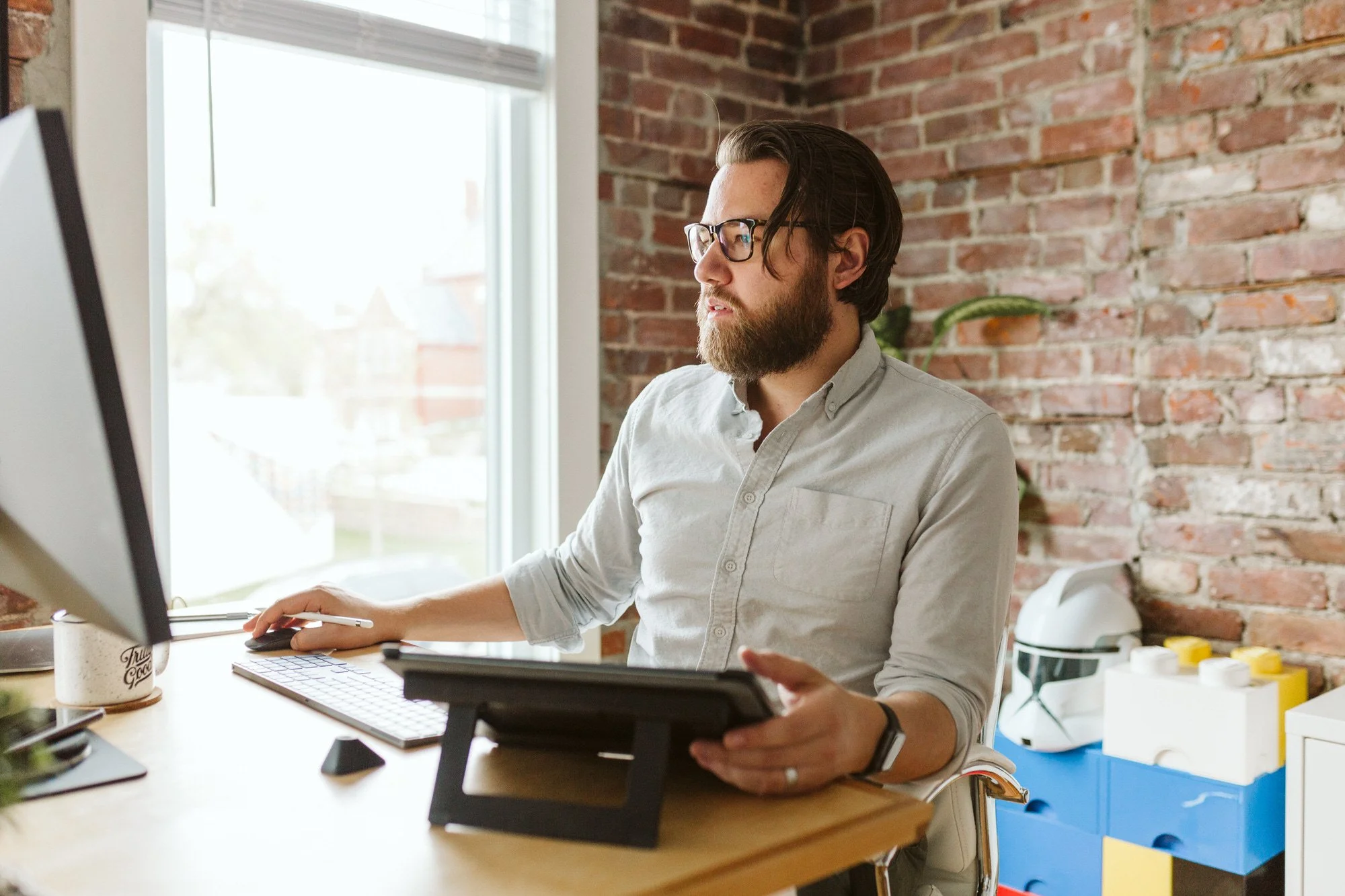 A man with glasses, a beard, and long dark hair sitting at a desk working on a computer with a graphics tablet, in a room with a brick wall and a window.