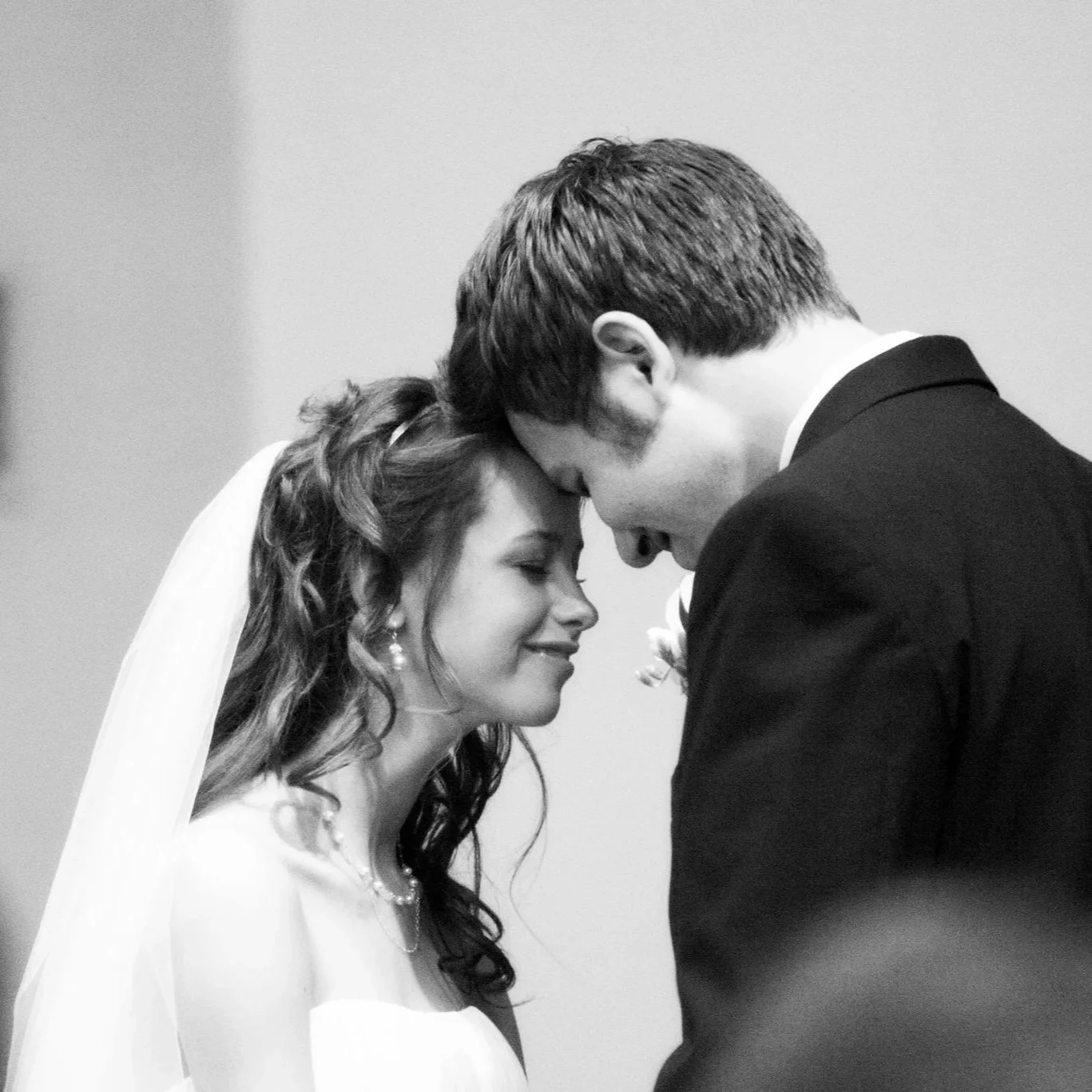 A bride and groom share a forehead kiss at their wedding, eyes closed, with a plain background.