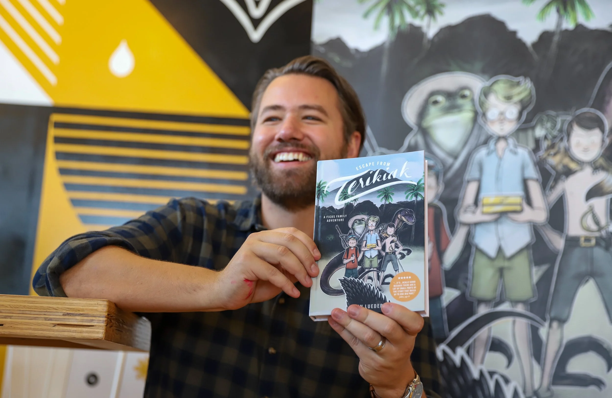 A man with a beard and plaid shirt smiling and holding a book titled 'Escape from Terikiak' in front of a colorful wall with cartoon characters and tropical trees.