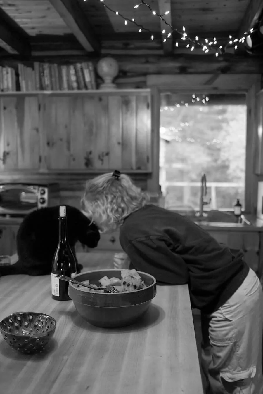 A person and a black cat leaning toward each other at a kitchen island with a large bowl of food, a bottle of wine, and a bowl with holes in it on the counter. The kitchen has wooden cabinets, a microwave, and string lights hanging from the ceiling.