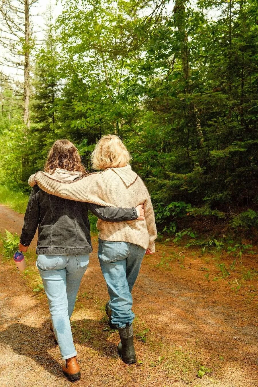 Two women walk together in a wooded area, one with her arm around the other's shoulders, as they walk along a dirt trail surrounded by green trees.