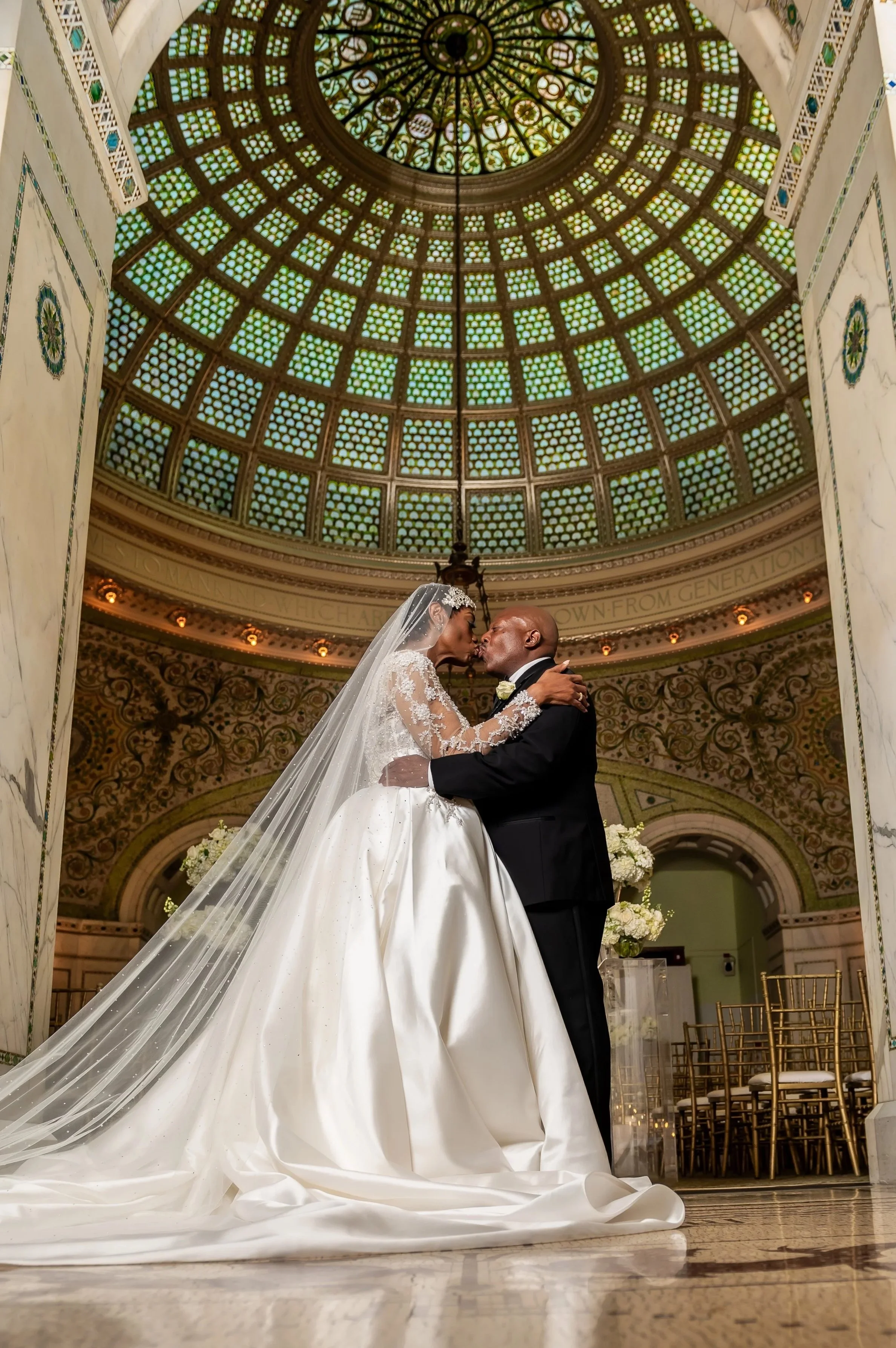 A bride and groom kissing during their wedding ceremony inside the Chicago Cultural Center with a stained glass dome ceiling.