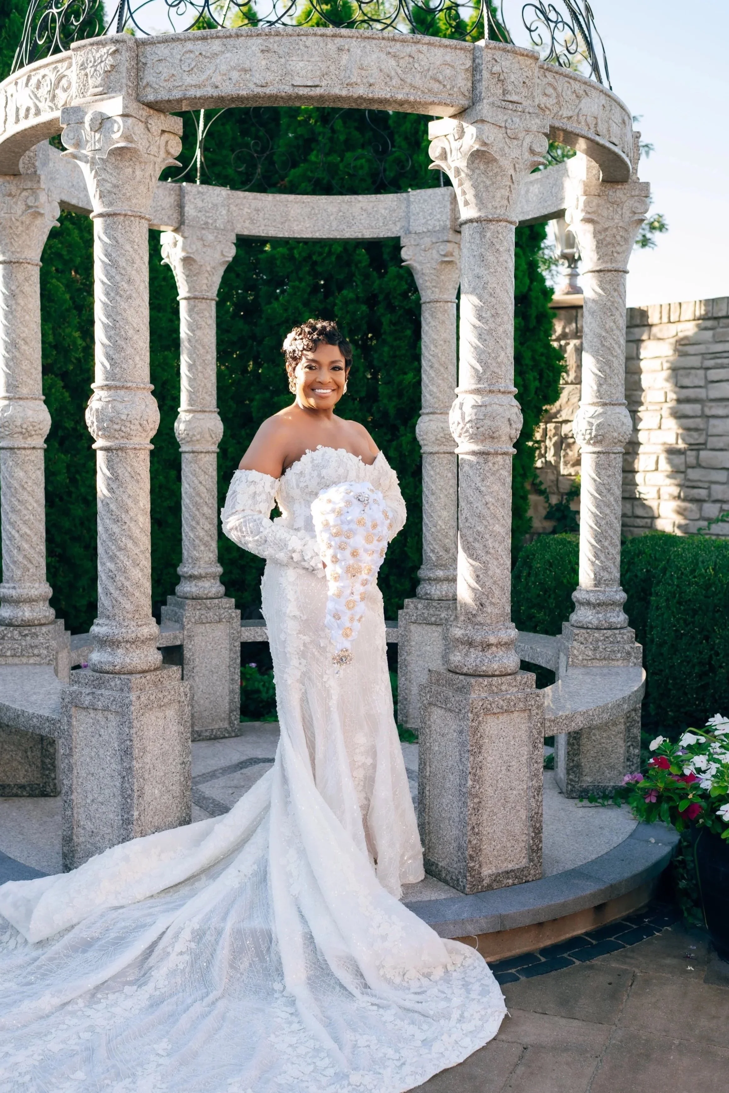 A smiling woman in a white wedding dress holding a bouquet of white flowers, standing under a stone gazebo with ornate ironwork details, in a garden with green bushes and flowering plants.