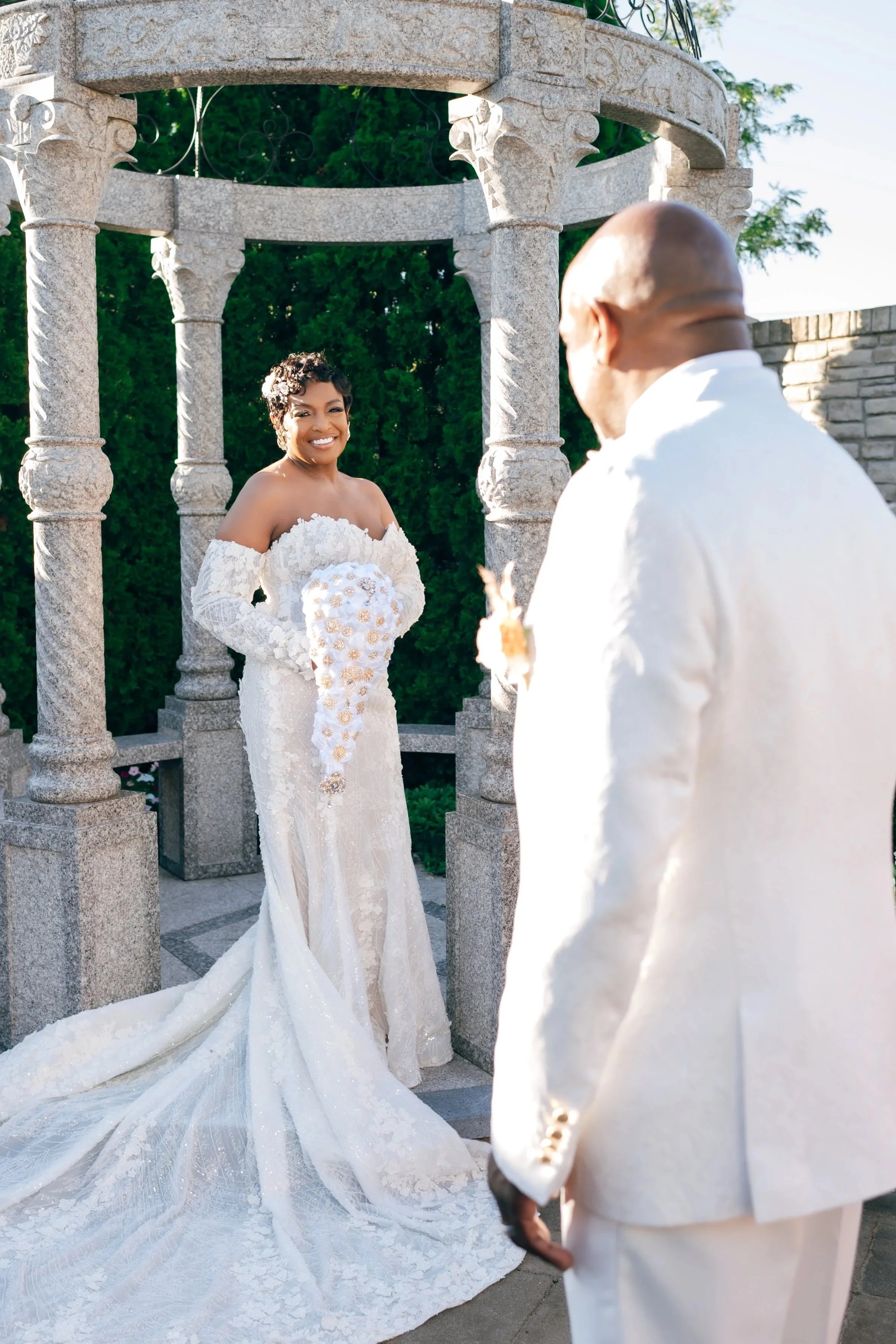 A bride in a white lace wedding dress holding a bouquet, smiling, standing in front of a stone gazebo, with the groom in a white suit facing her, outdoors with greenery and a stone wall in the background.
