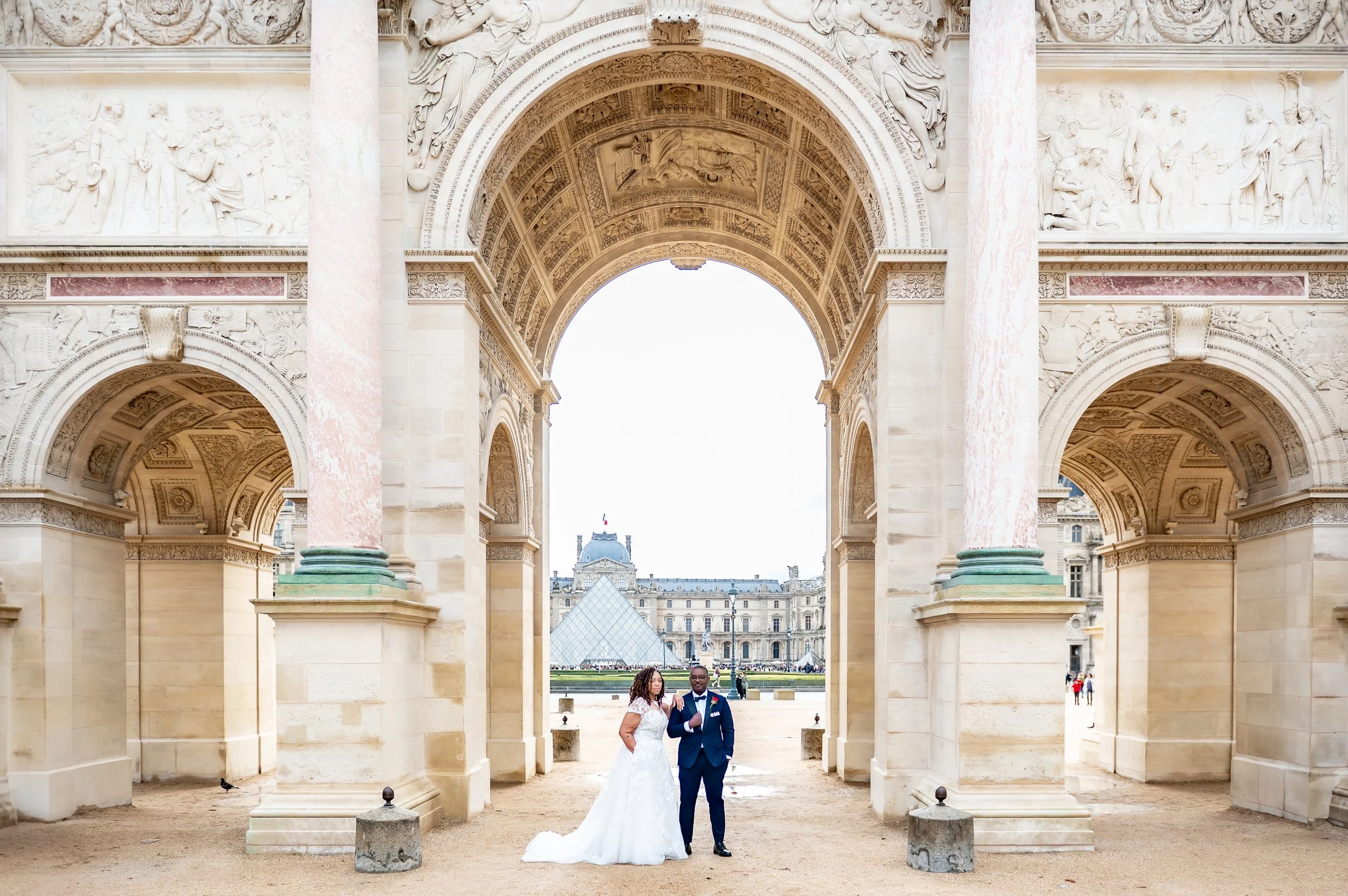A bride and groom in wedding attire posing under a classical stone archway in front of the Louvre Pyramid in Paris, with historic buildings and tourists in the background.