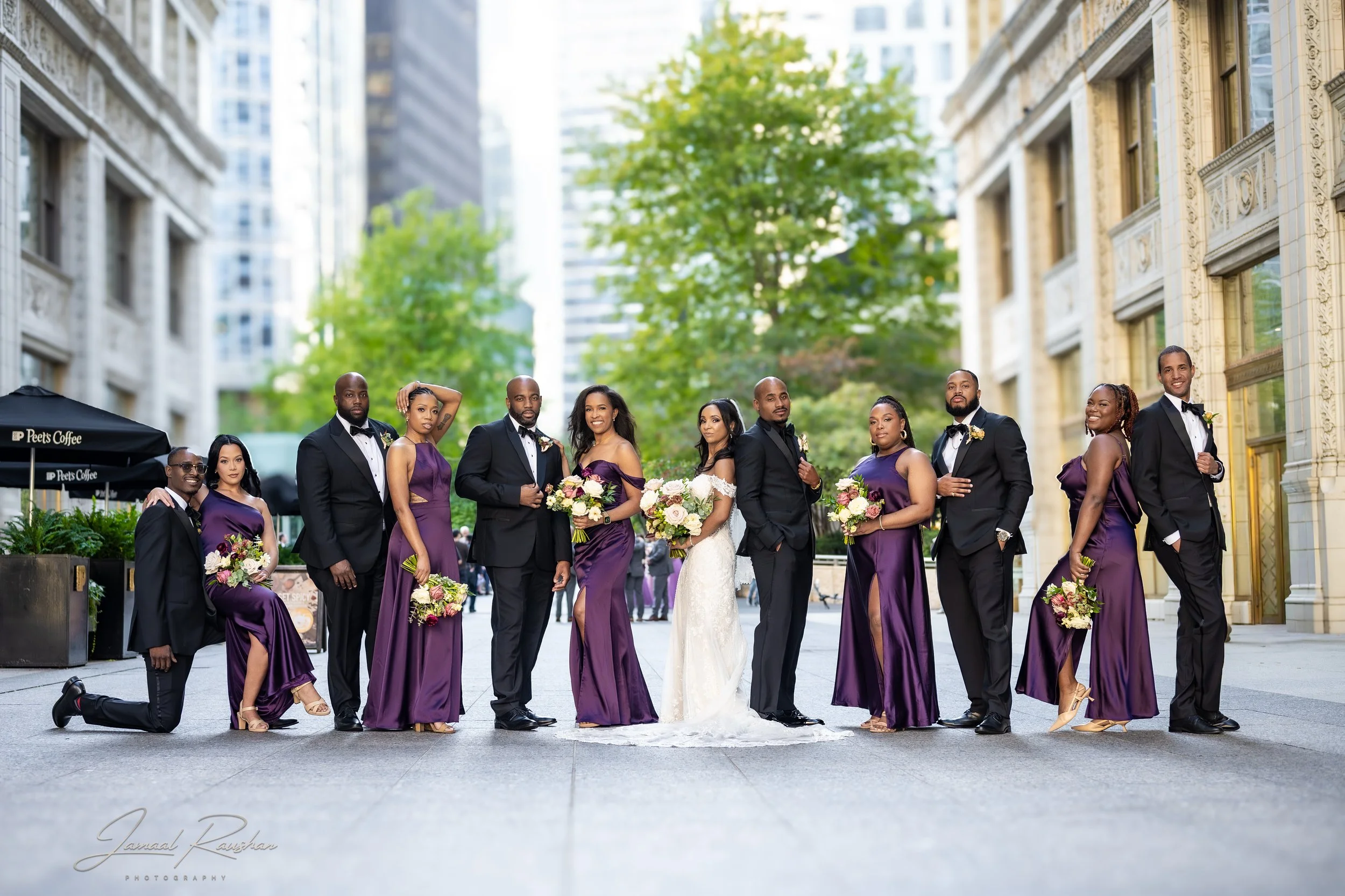 A wedding party posing for a photo outdoors near the Wrigley building in Chicago and trees in the background. The women are wearing purple dresses, and the men are in black tuxedos.