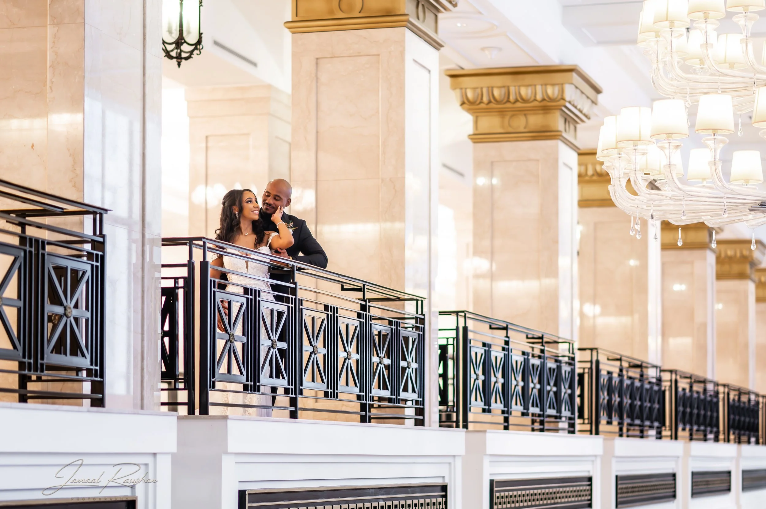 A smiling couple dressed in formal attire, standing on a balcony railing in a grand, elegant interior with chandeliers and large columns. The JW Marriott hotel in Chicago