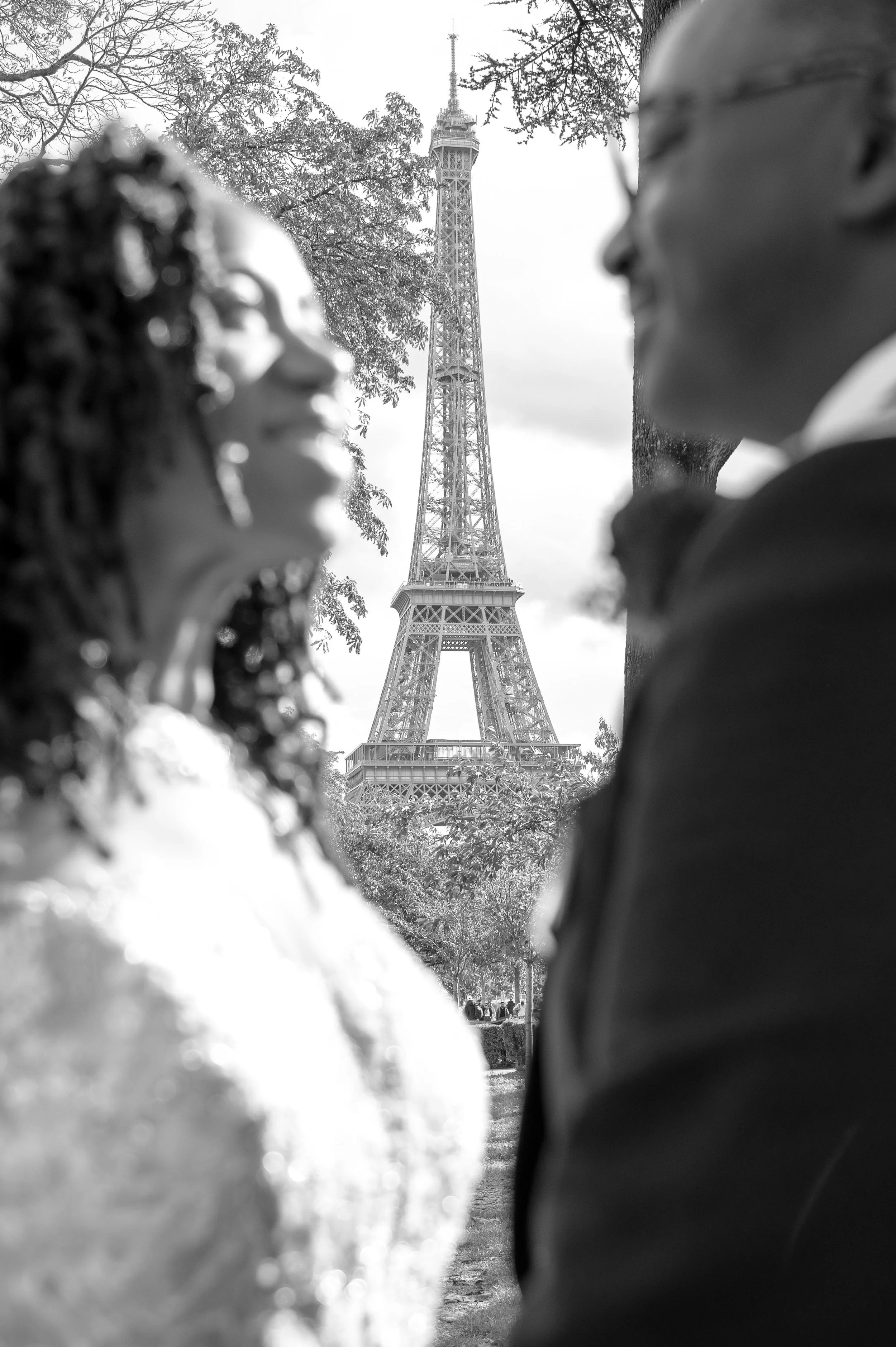 A black and white photograph of the Eiffel Tower seen through silhouettes of two people, a woman and a man, facing each other, with trees in the background.