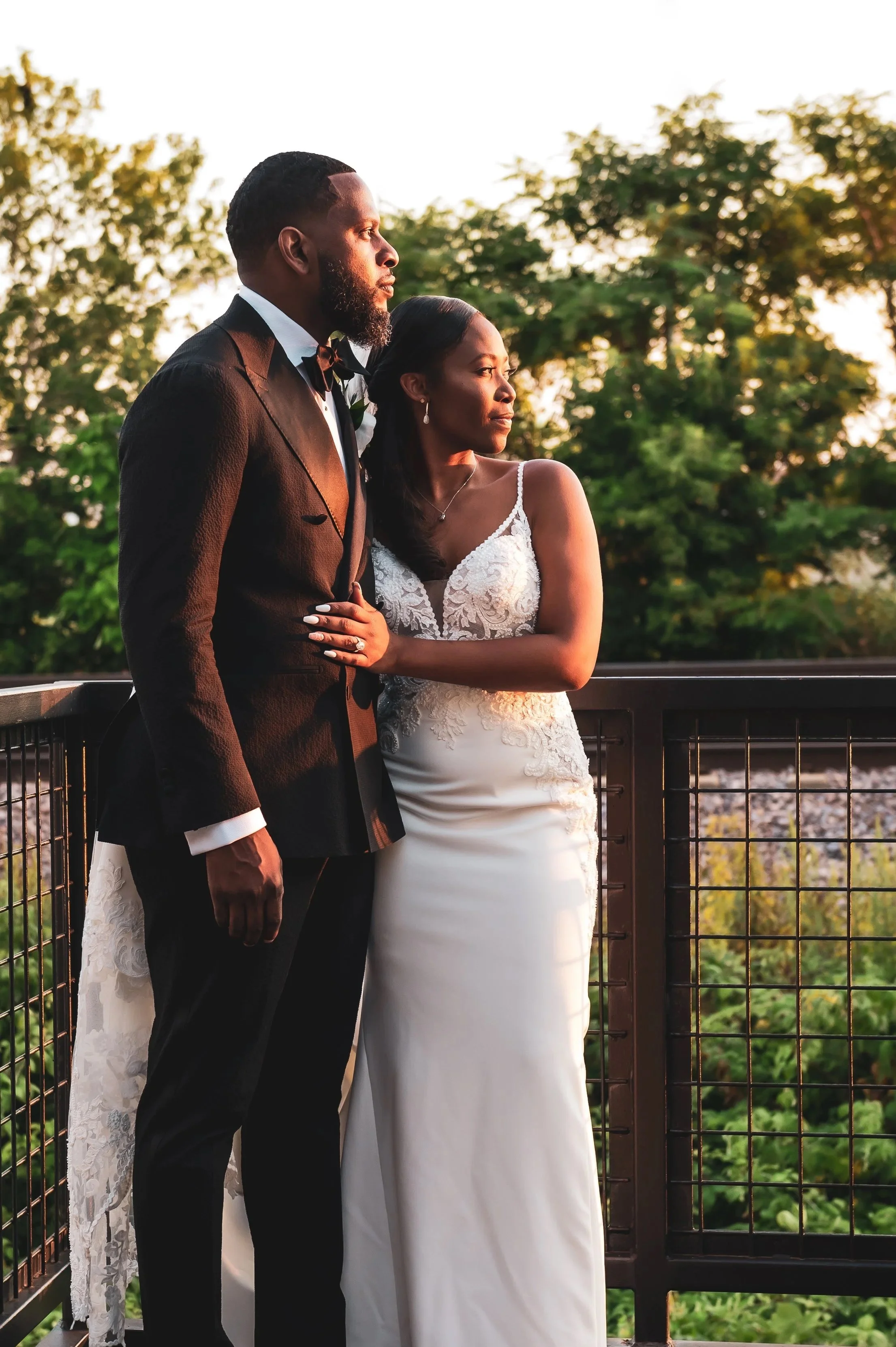 A bride and groom dressed in wedding attire on a bridge with trees and greenery in the background during sunset.