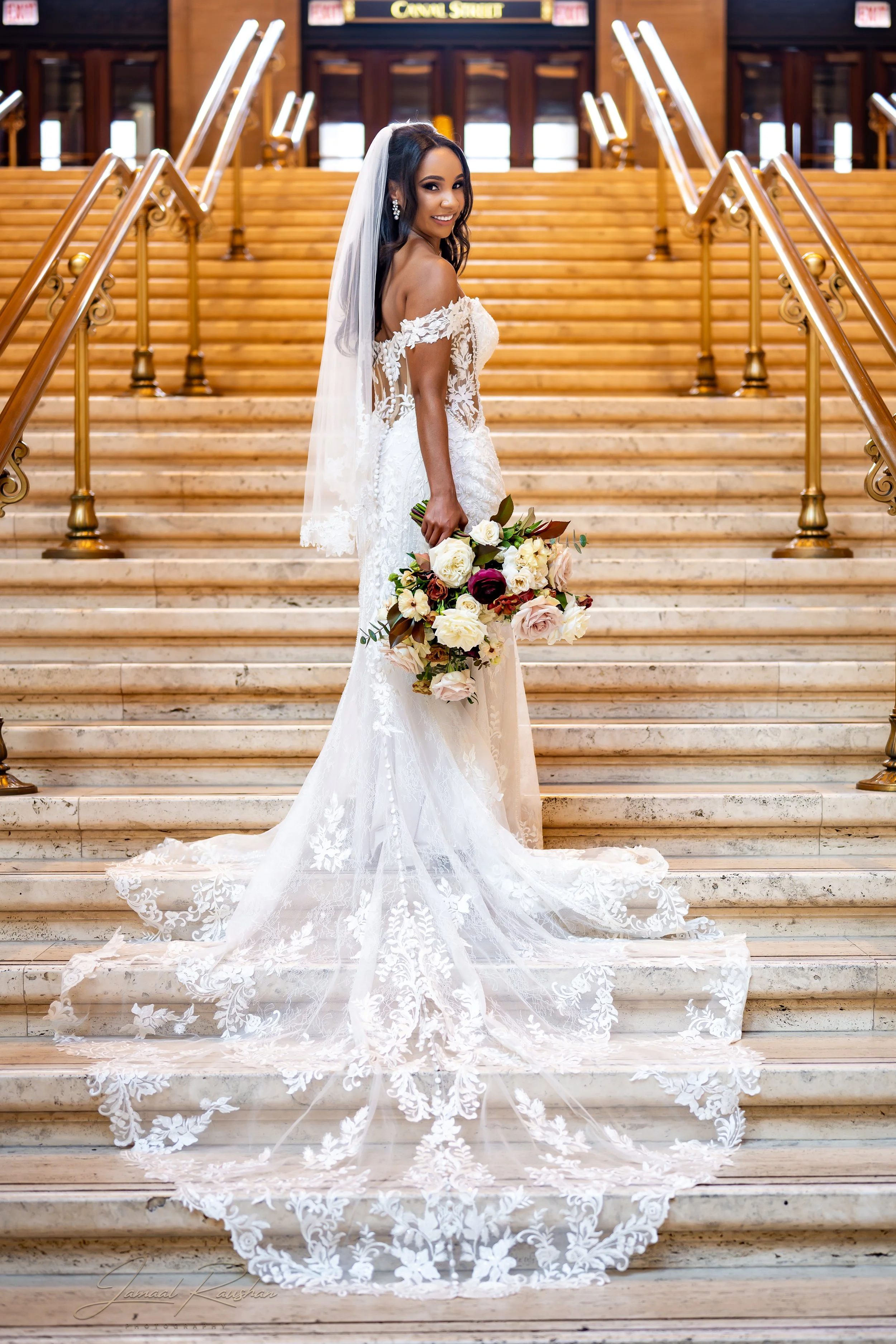 A bride in a white lace wedding dress standing on a marble staircase at Union Station in Chicago holding a bouquet of flowers, smiling. The staircase has wooden handrails and a background of wood-paneled walls and doorways.