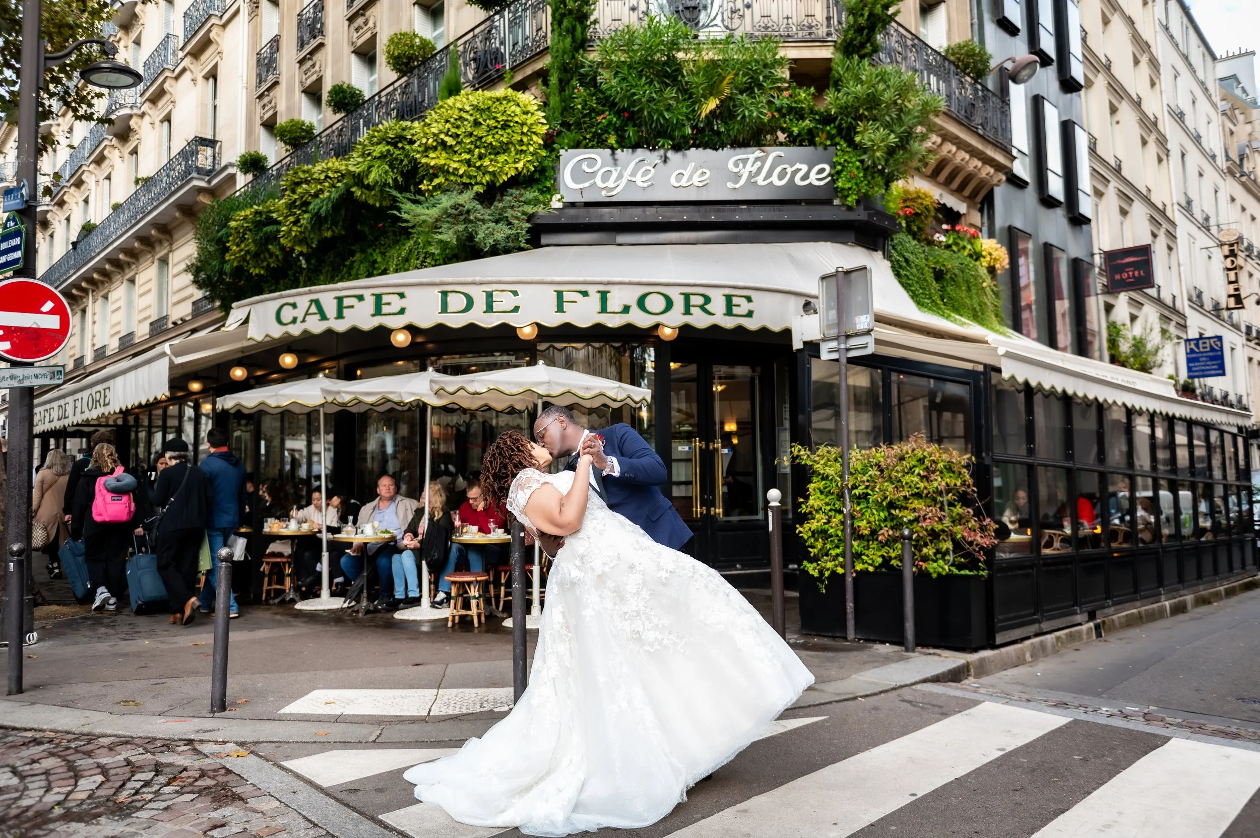 A bride and groom kiss outside a Parisian café named Cafe de Flore, with a crowd watching them. The café has a black awning and green plants on the second-floor balcony.